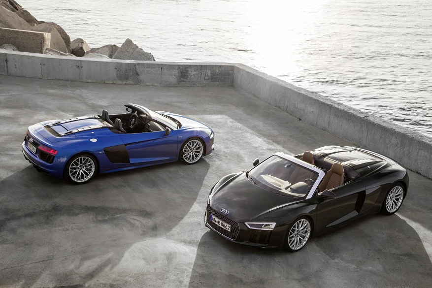 Zwei Audi R8 Cabrios geparkt am Meer, einer blau, der andere schwarz. Beide mit geöffnetem Verdeck auf einer Betonfläche.