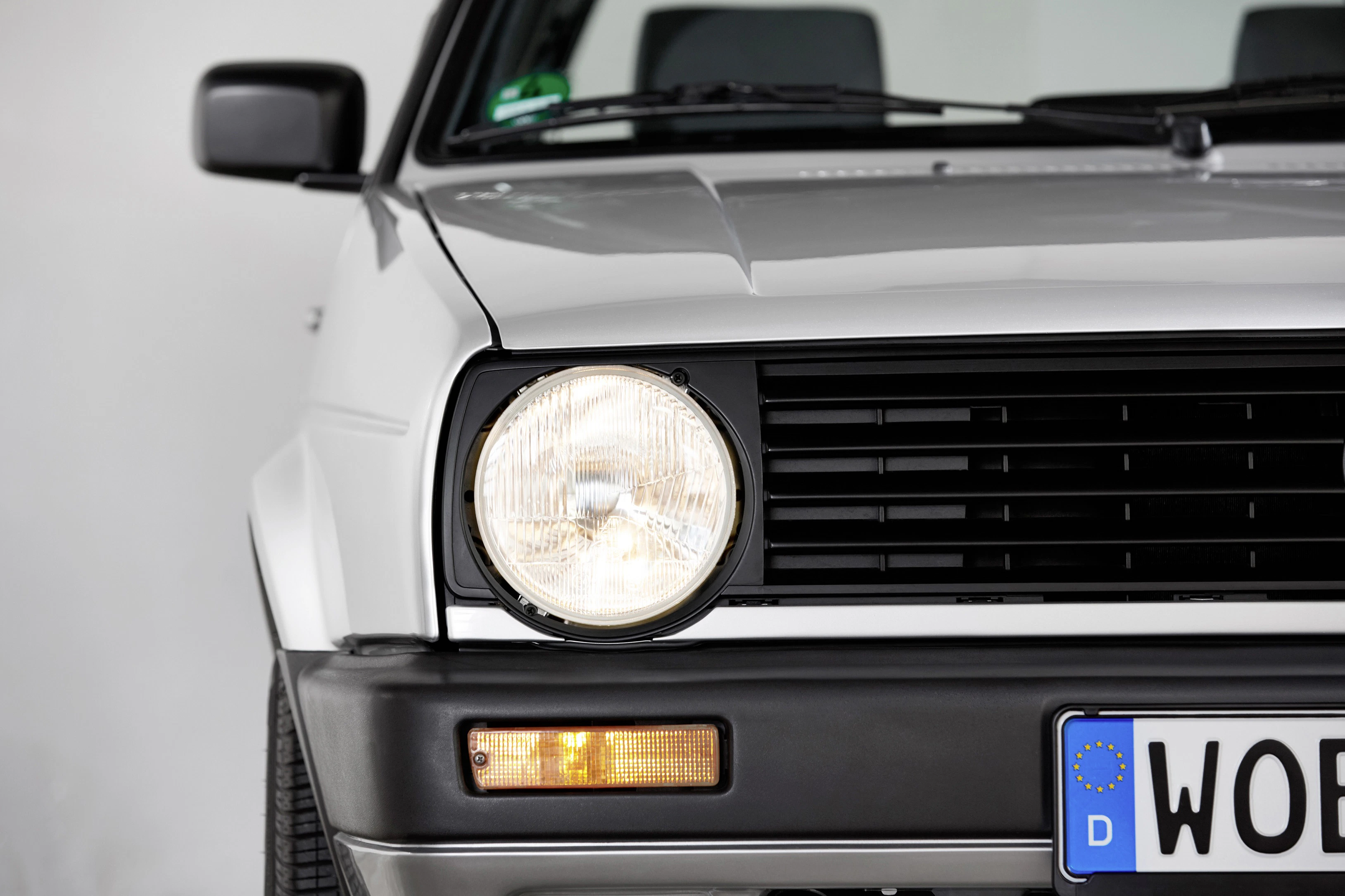 Nahaufnahme eines weißen Autos, Fokus auf Frontscheinwerfer und Nummernschild. Modernes, minimales Design mit spiegelndem Lack.