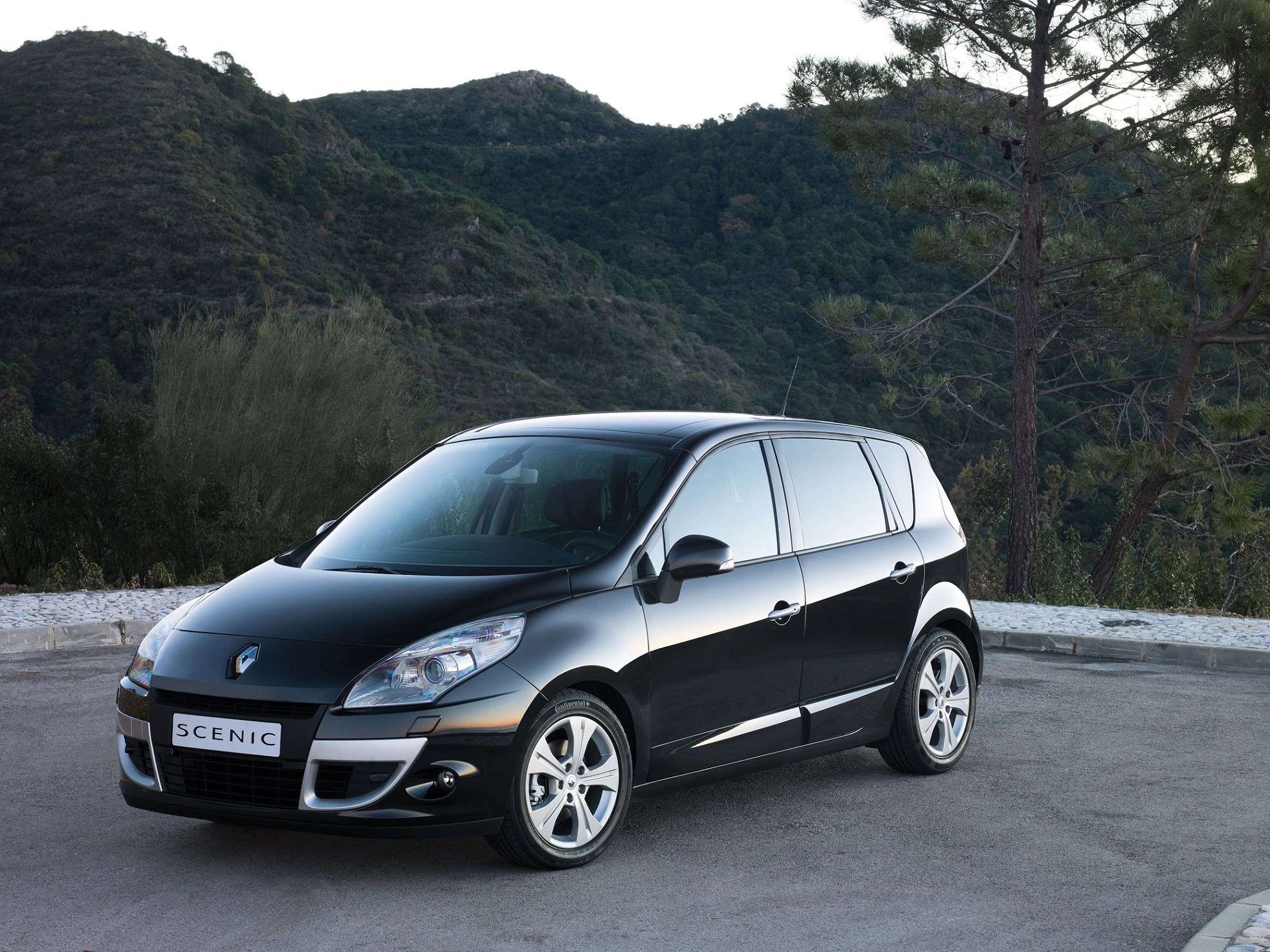 Schwarzer Renault Scenic steht auf einem Parkplatz vor bewaldeten Hügeln und Bäumen unter klarem Himmel.