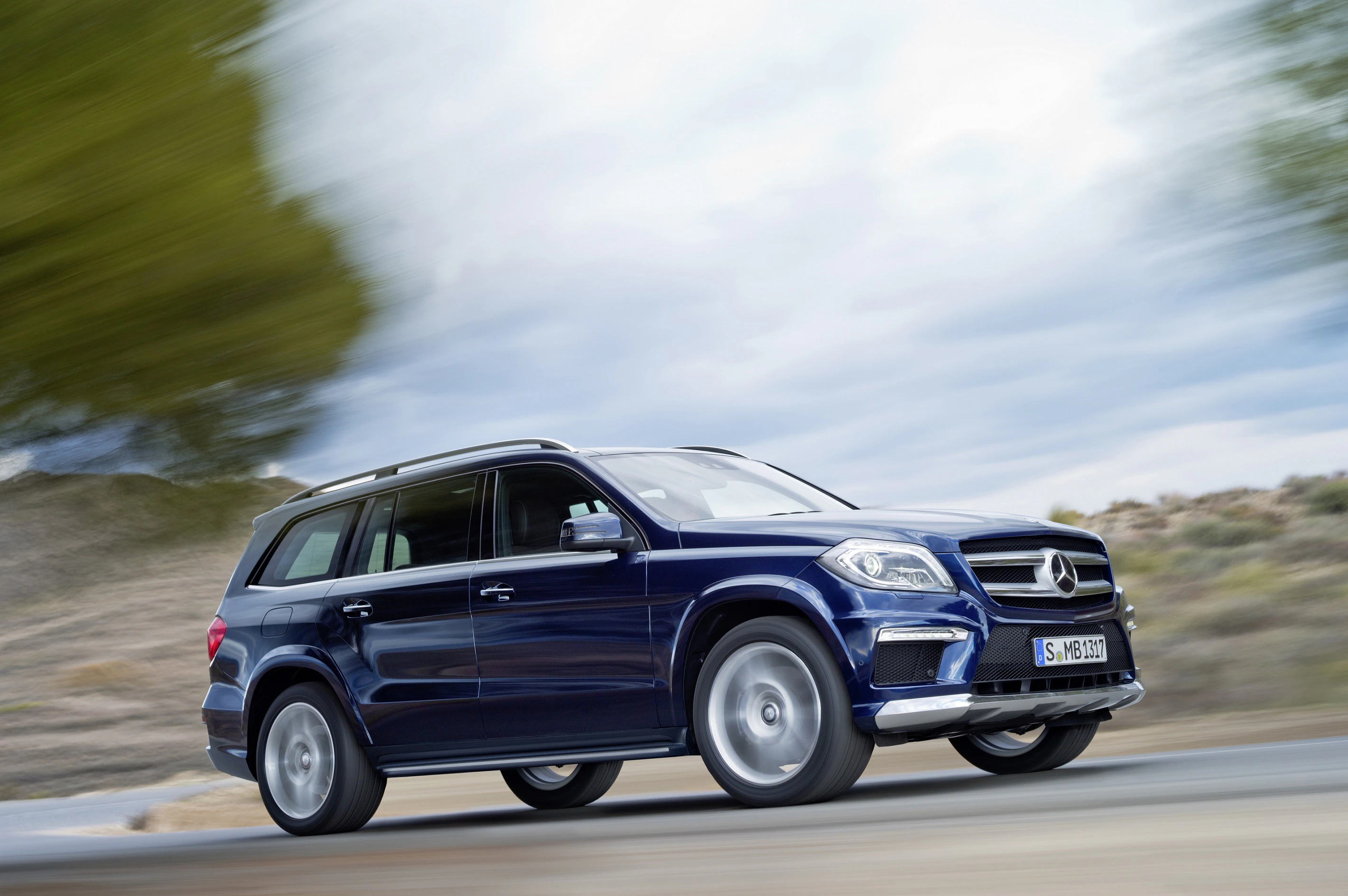 Blauer Mercedes-Geländewagen fährt dynamisch auf einer Straße, umgeben von verschwommenen Bäumen und bewölktem Himmel im Hintergrund.