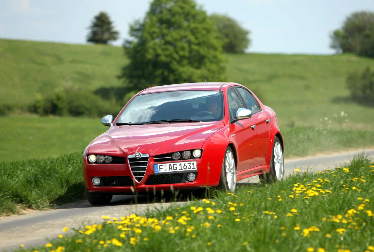 Roter Alfa Romeo fährt auf schmalem Landweg durch grüne Wiesen mit gelben Blumen, im Hintergrund Bäume und blauer Himmel.
