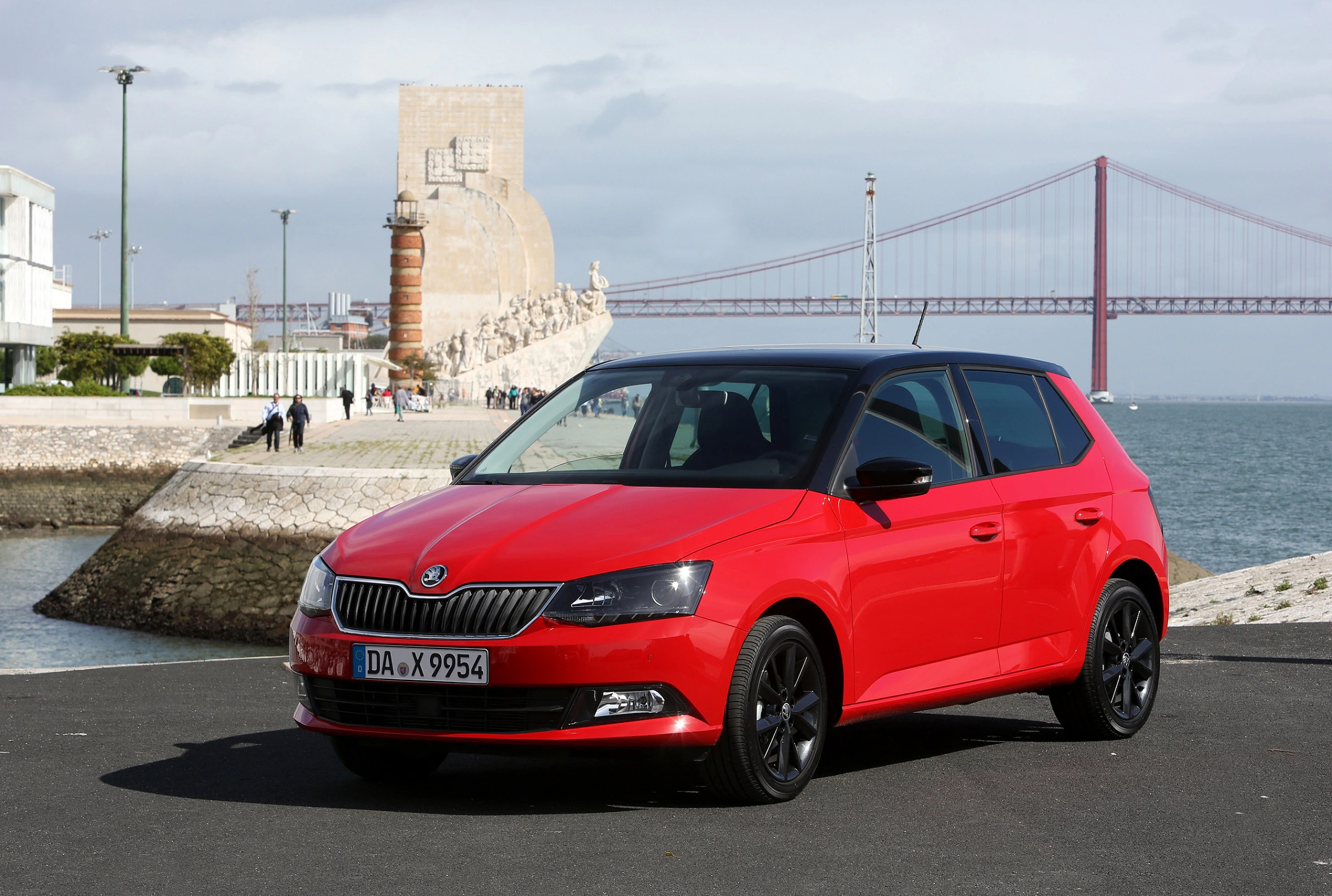 Rotes Auto vor dem Entdeckerdenkmal und der Brücke des 25. April in Lissabon, bei bewölktem Himmel.