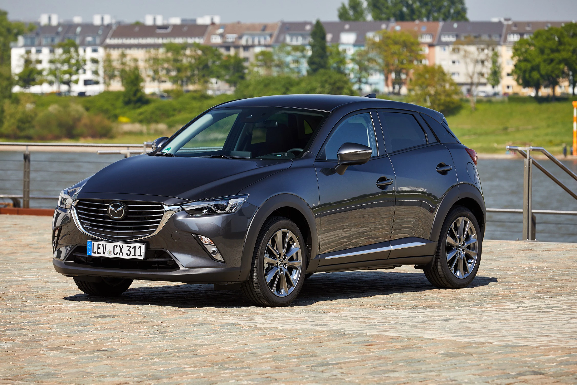 Grauer Mazda CX-3 SUV steht auf einer gepflasterten Fläche, im Hintergrund ein Fluss und Stadthäuser.