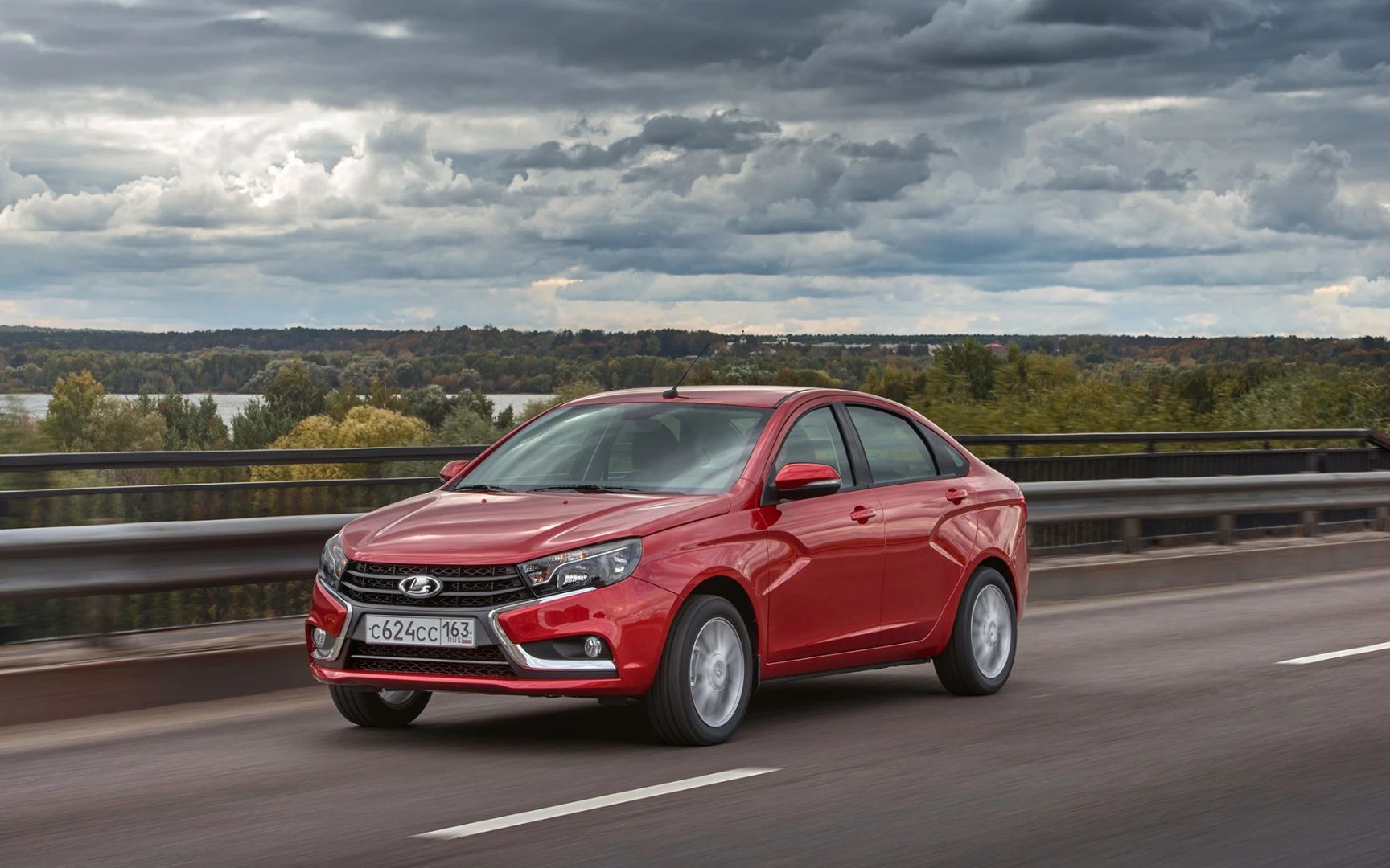 Rotes Auto fährt auf einer Landstraße, im Hintergrund eine bewaldete Landschaft und ein bedeckter Himmel.