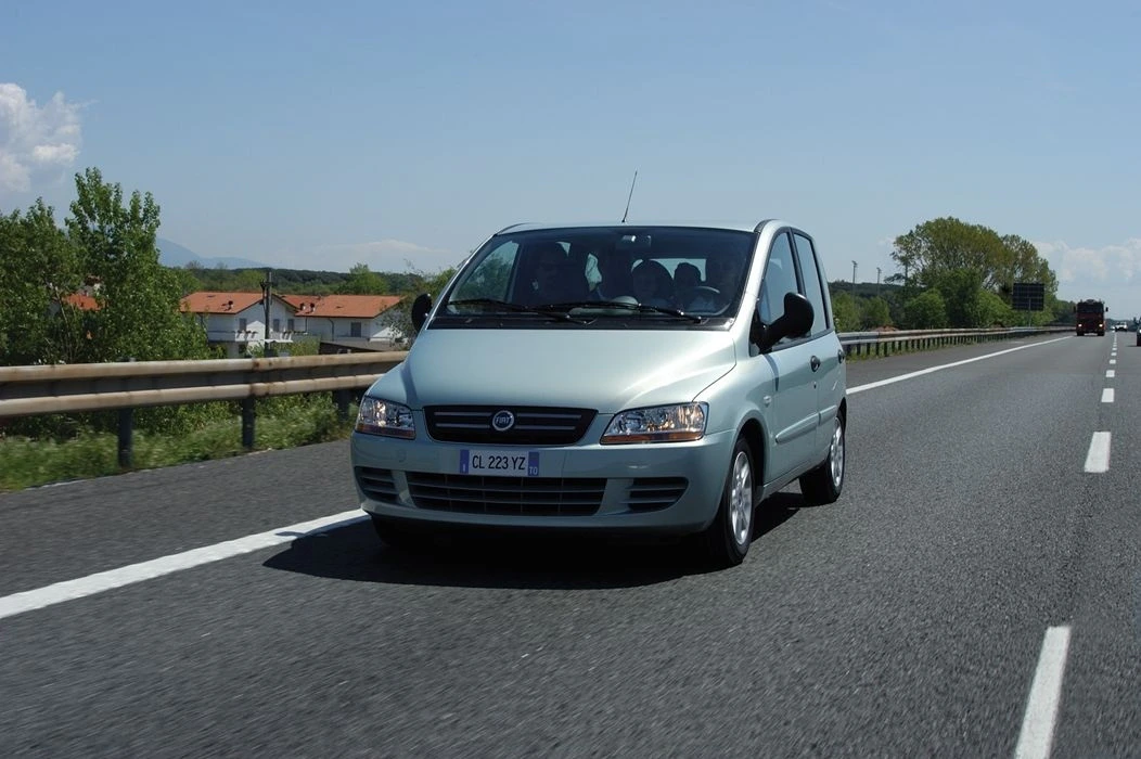 Silberner Fiat Multipla fährt auf einer Autobahn, sonniges Wetter, Bäume und Häuser im Hintergrund.