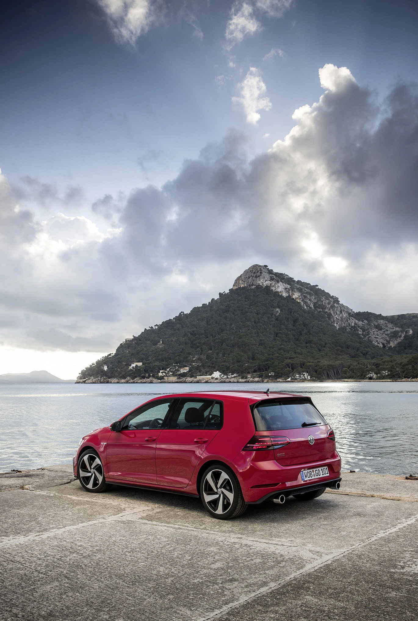 Roter VW Golf steht auf einem Parkplatz am Meer, im Hintergrund ein bewaldeter Berg unter bewölktem Himmel.