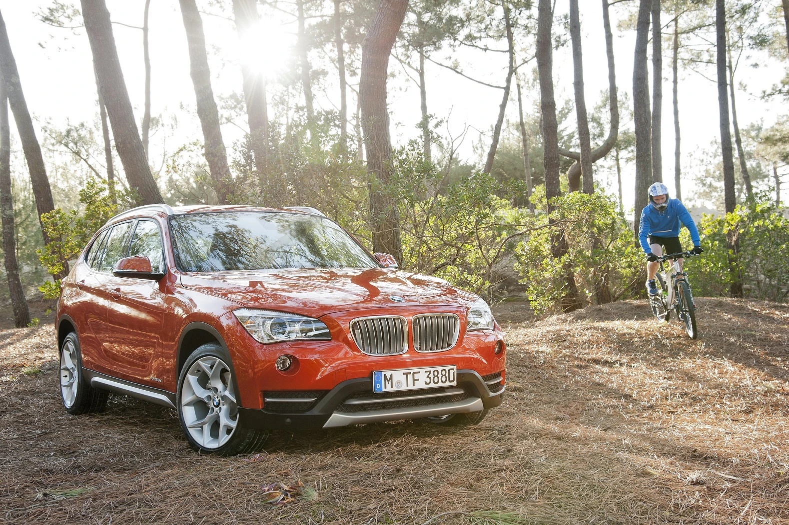 Roter BMW-SUV parkt im Wald auf einem Nadelboden. Ein Radfahrer in blauer Kleidung fährt neben dem Auto durch die Bäume.