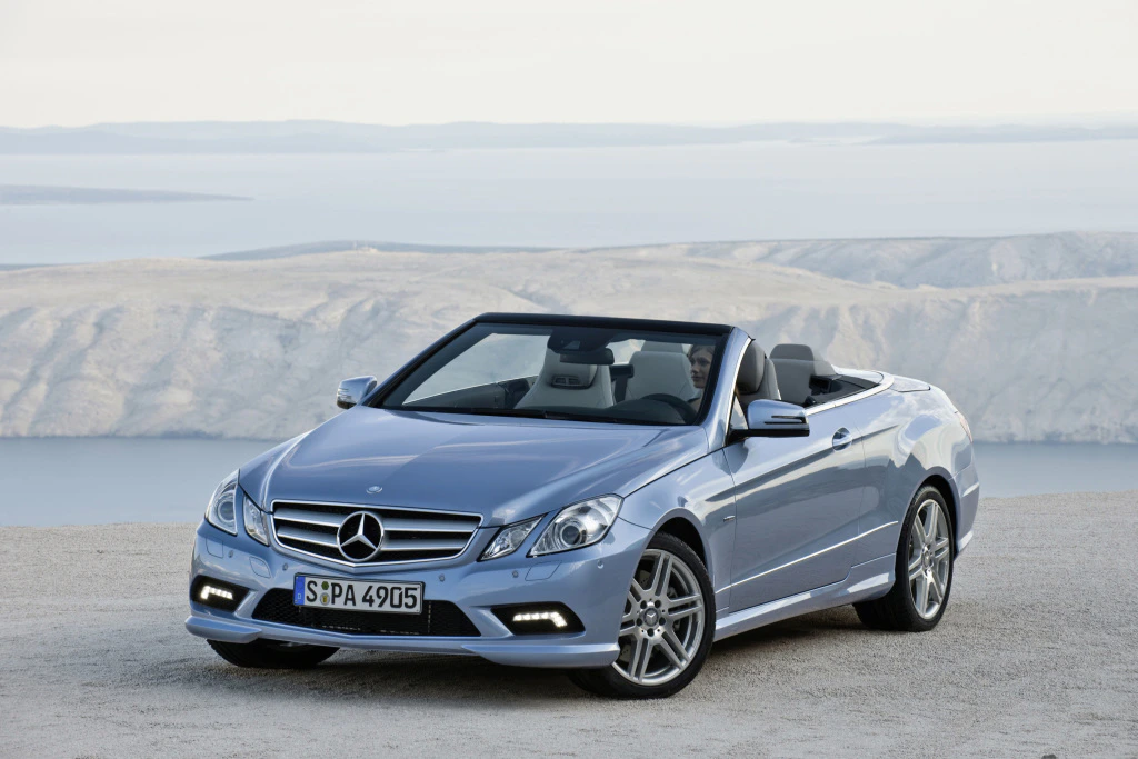 Silbernes Mercedes-Benz Cabriolet parkt auf sandigem Gelände, im Hintergrund eine weite Landschaft mit Wasser und Hügeln.