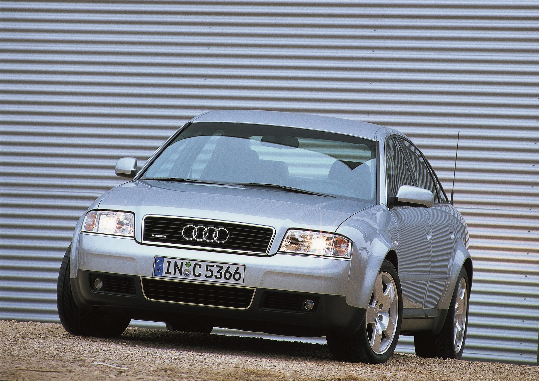 Silberner Audi vor einem wellenförmigen, metallischen Hintergrund. Sicht von vorne links, Scheinwerfer eingeschaltet.
