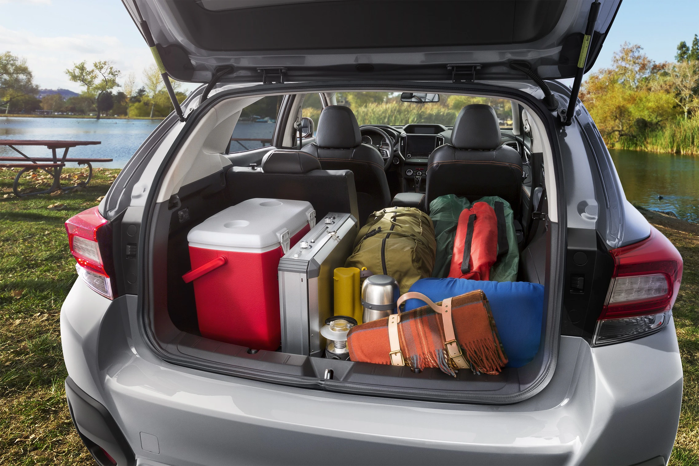 Ein geöffneter Kofferraum eines Autos am See, gefüllt mit Campingausrüstung wie Kühlbox, Rucksäcke und Isomatten.