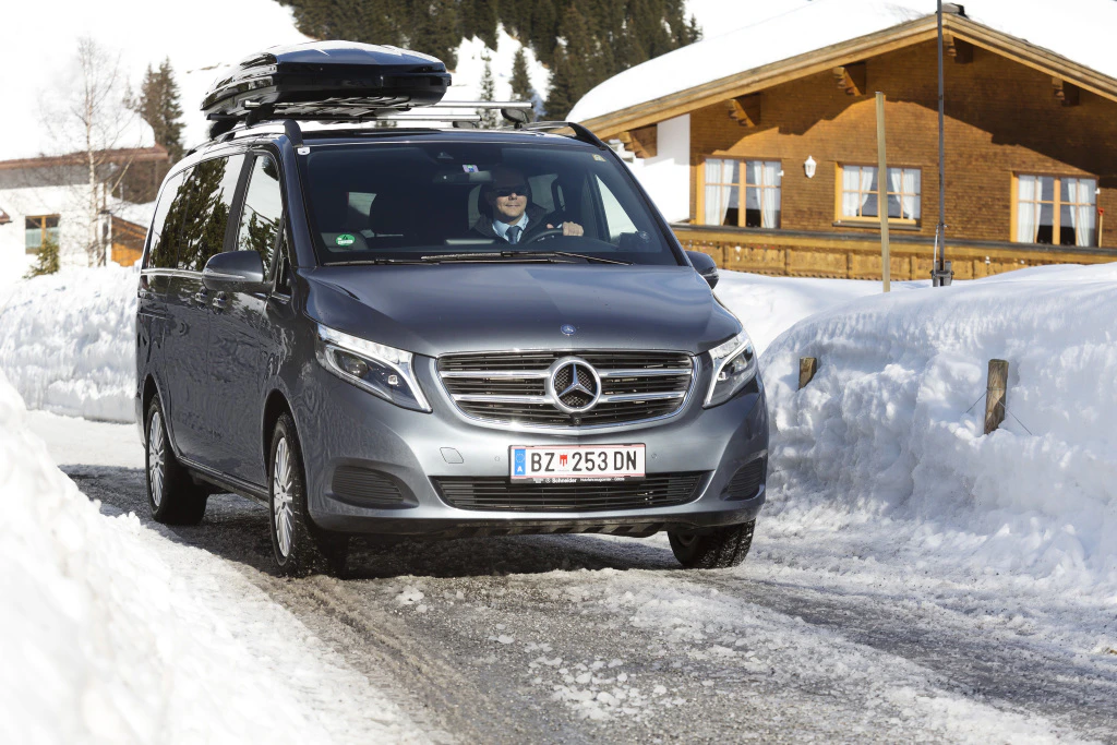Silberner Mercedes-Van auf schneebedeckter Straße, mit Dachbox und einer winterlichen Hütte im Hintergrund.