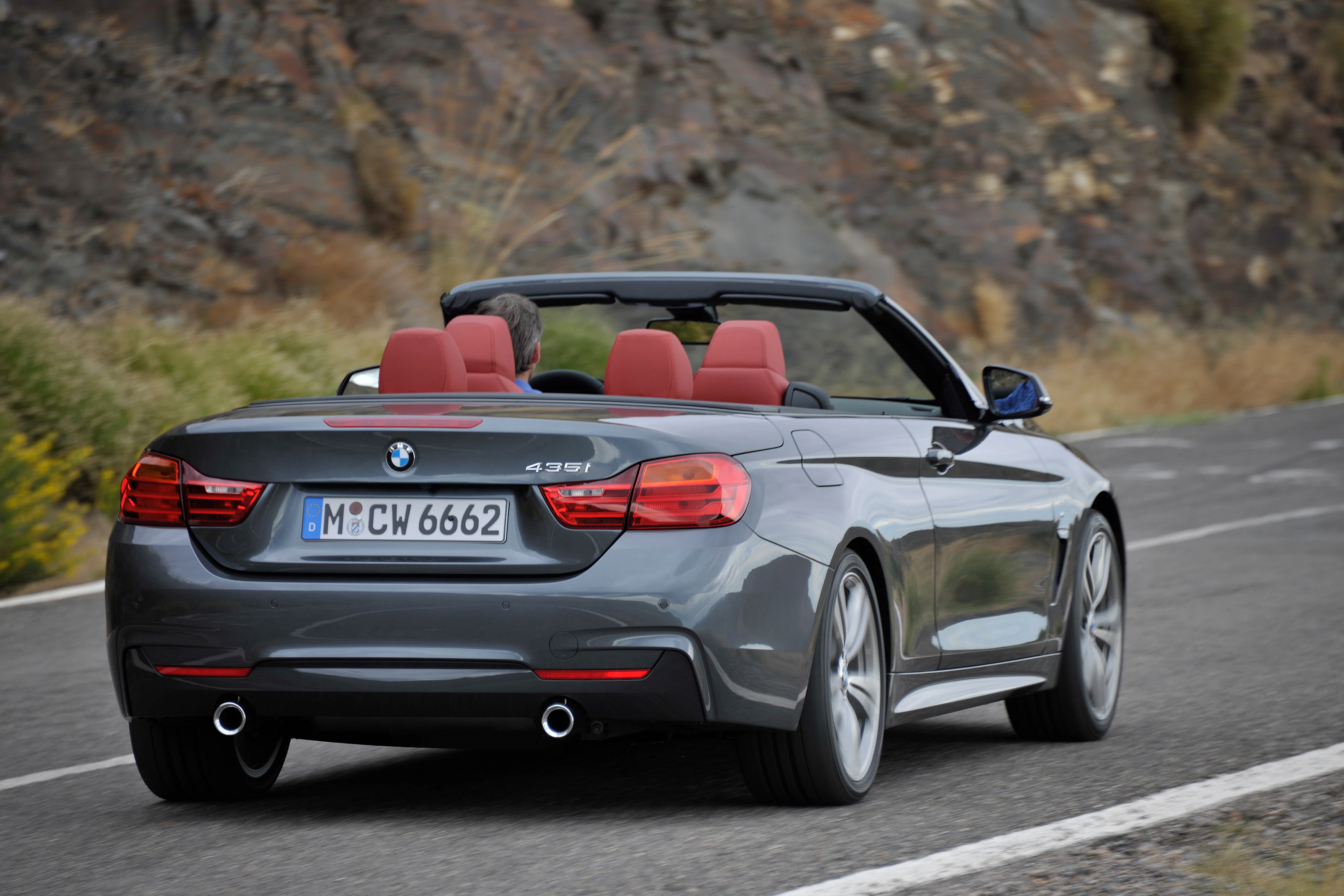Grau-metallic BMW Cabrio fährt auf einer Landstraße. Das Verdeck ist geöffnet, rote Ledersitze sind sichtbar, umgeben von Felsen und Vegetation.