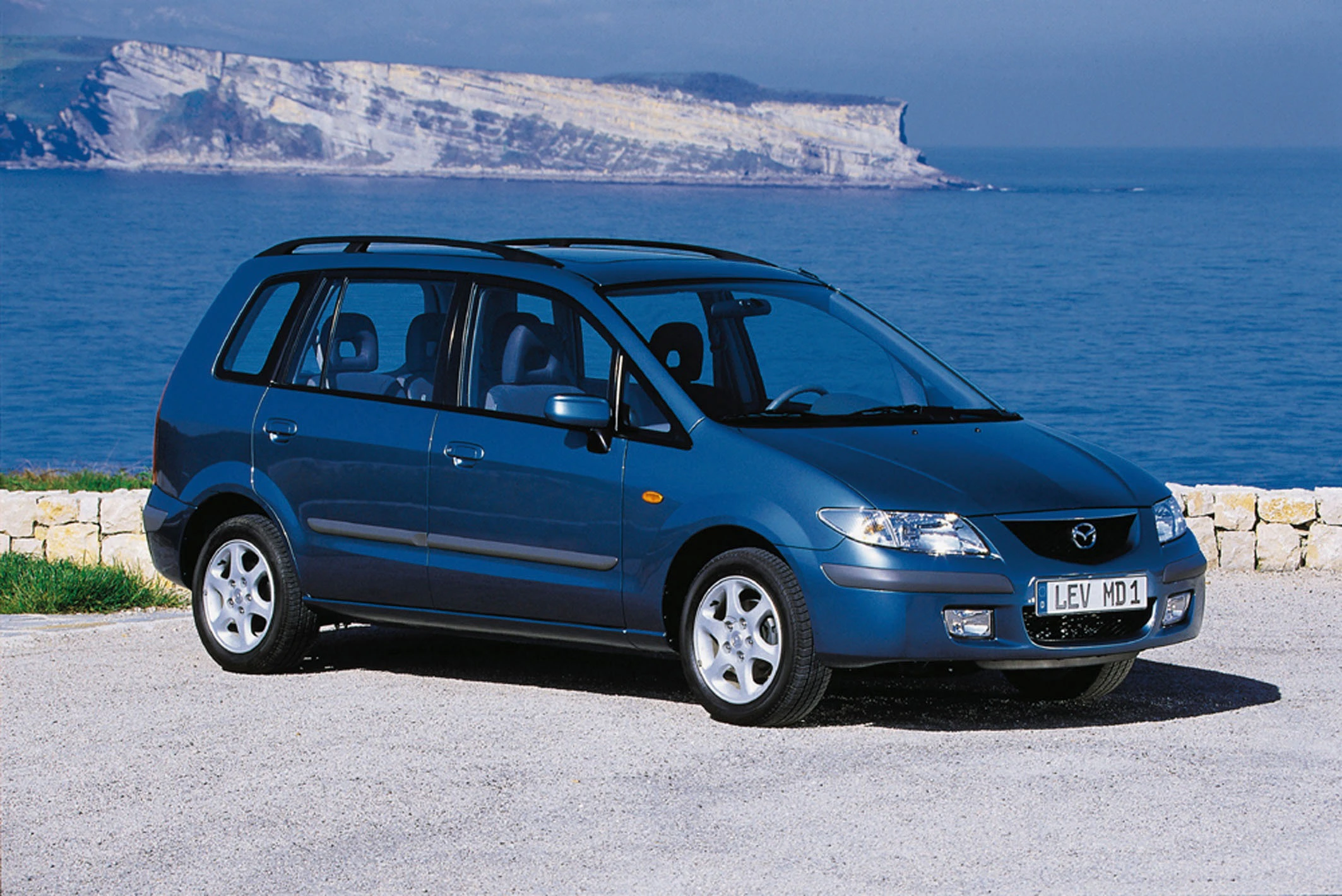 Blauer Mazda-Van parkt vor blauem Meer und felsiger Küste; sonniger Himmel.