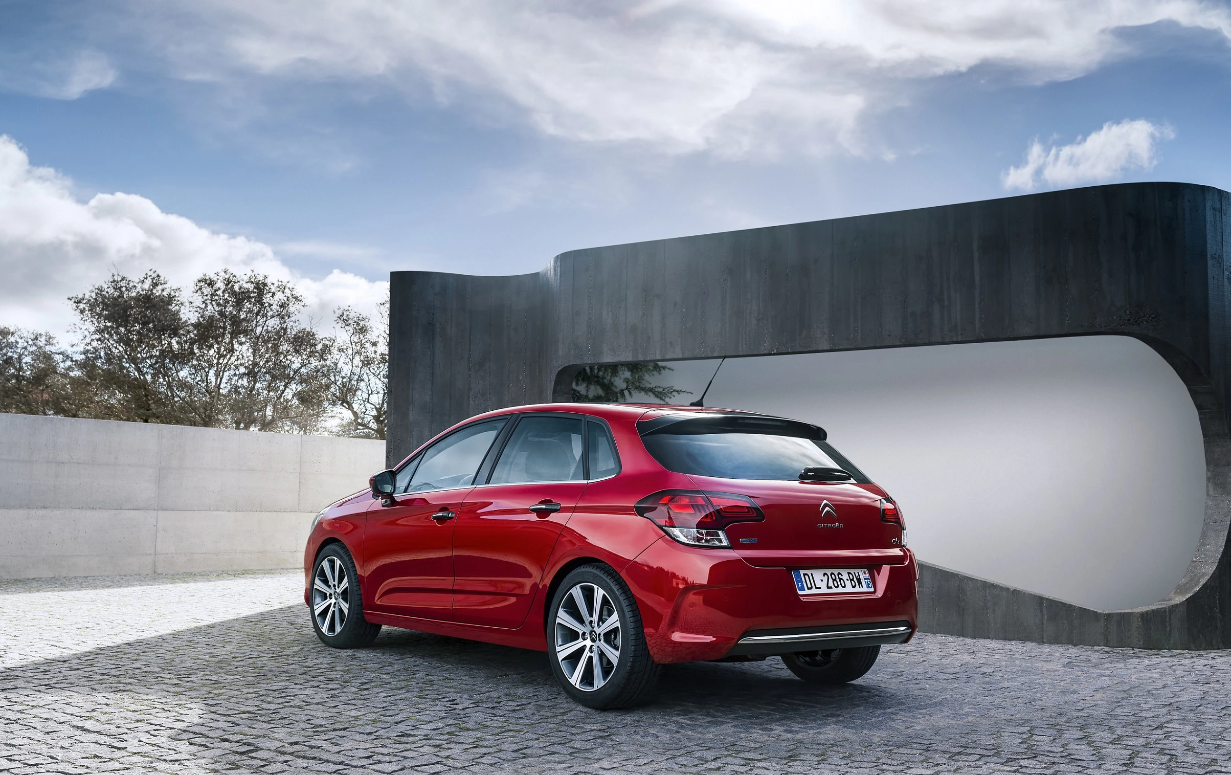 Roter Citroën C4 parkt auf gepflastertem Platz vor modernem, minimalistischen Gebäude unter bewölktem Himmel.