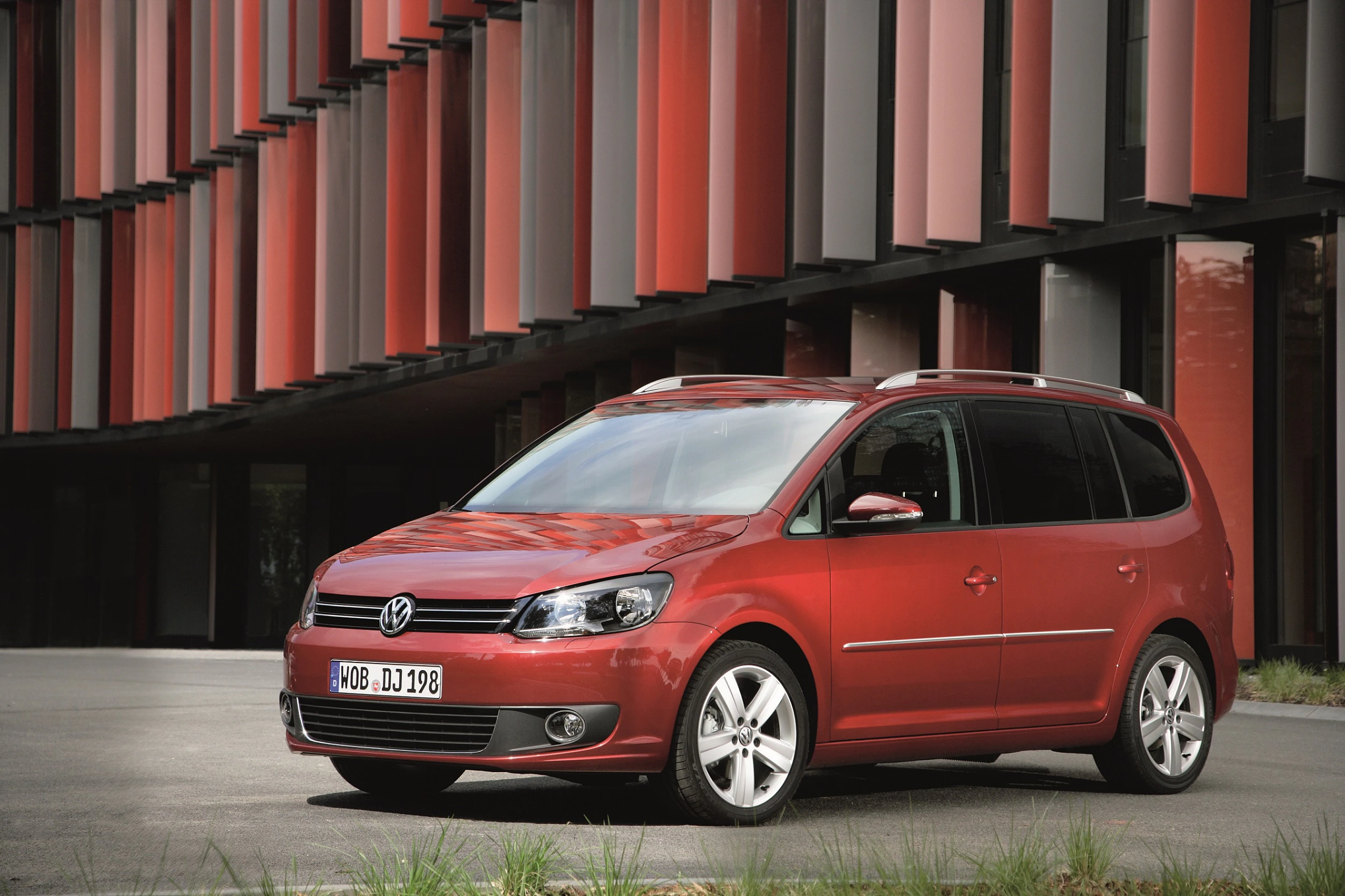 Roter Volkswagen Touran steht vor einem modernen Gebäude mit großen Glas- und Metallflächen.