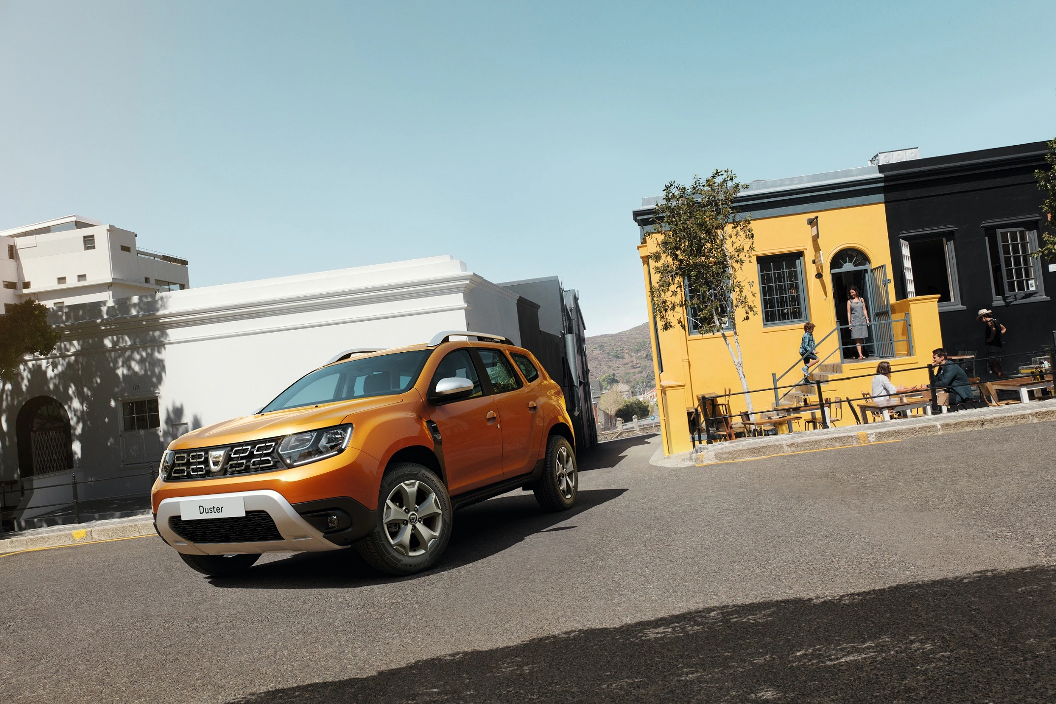Orangefarbenes SUV parkt auf einer Straße zwischen bunten Gebäuden. Einige Menschen sitzen draußen und genießen den sonnigen Tag.