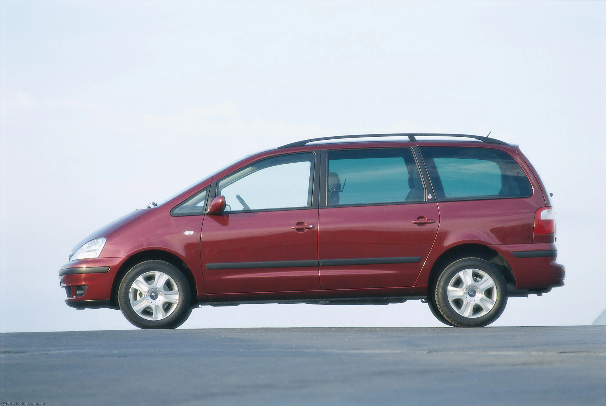 Roter Minivan mit fünf Türen, seitlich gegen einen wolkigen Himmel auf einer asphaltierten Fläche geparkt.