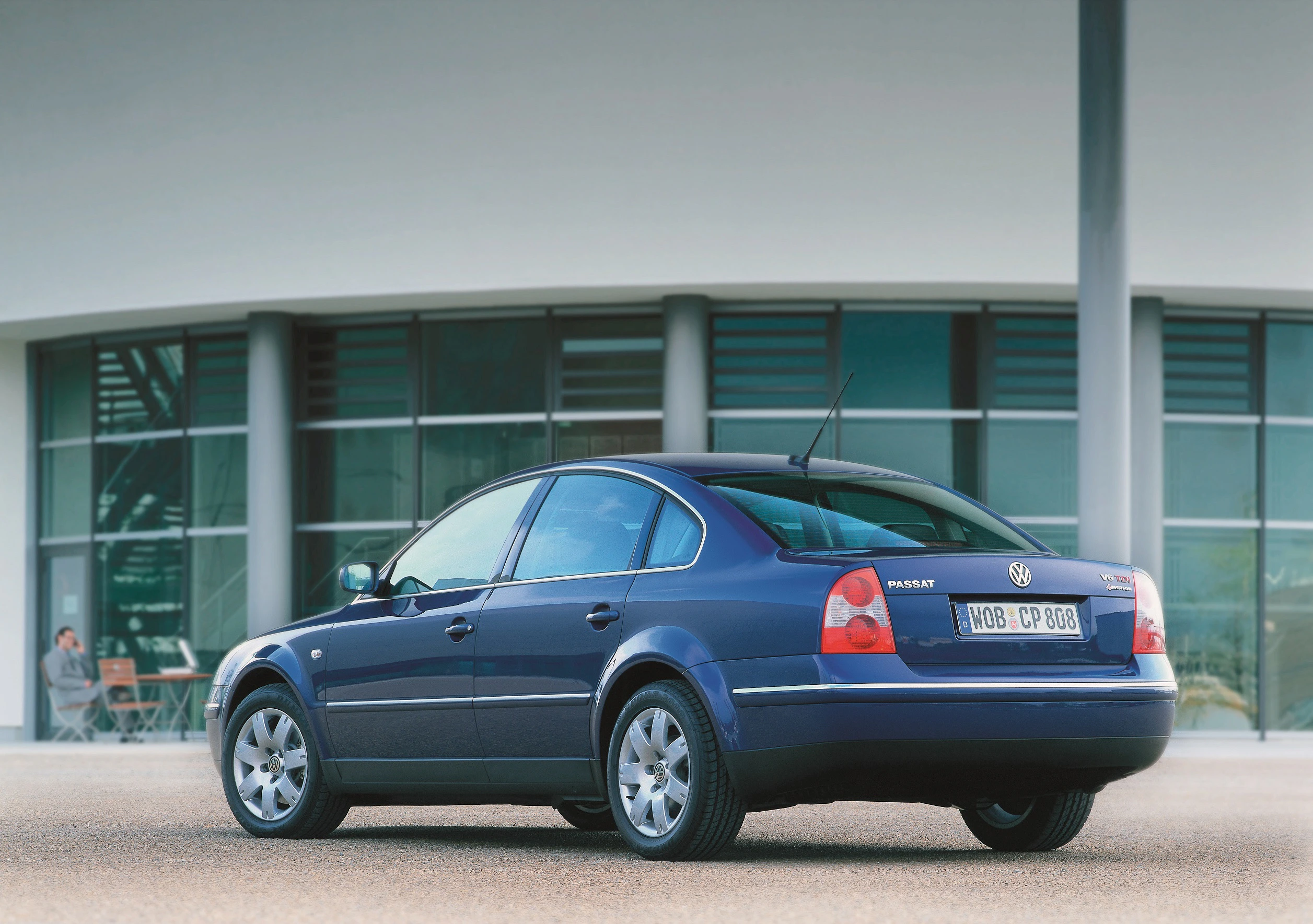 Blauer Volkswagen Passat Limousine steht vor einem modernen Gebäude mit großen Fenstern. Rückansicht des Autos auf leerem Parkplatz.