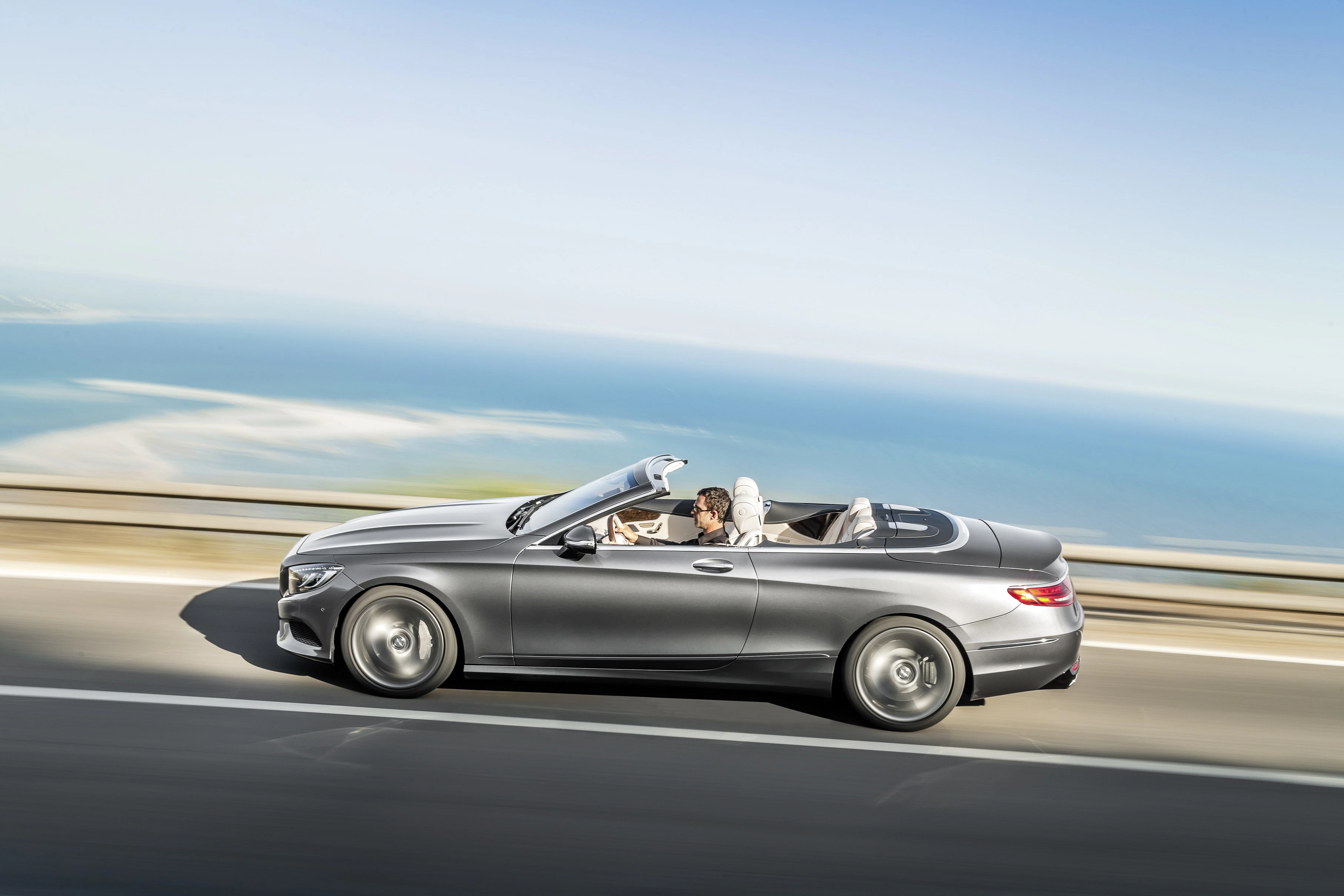 Silbernes Cabriolet fährt an Küste entlang; Fahrer genießt Ausblick aufs Meer unter klarem Himmel.