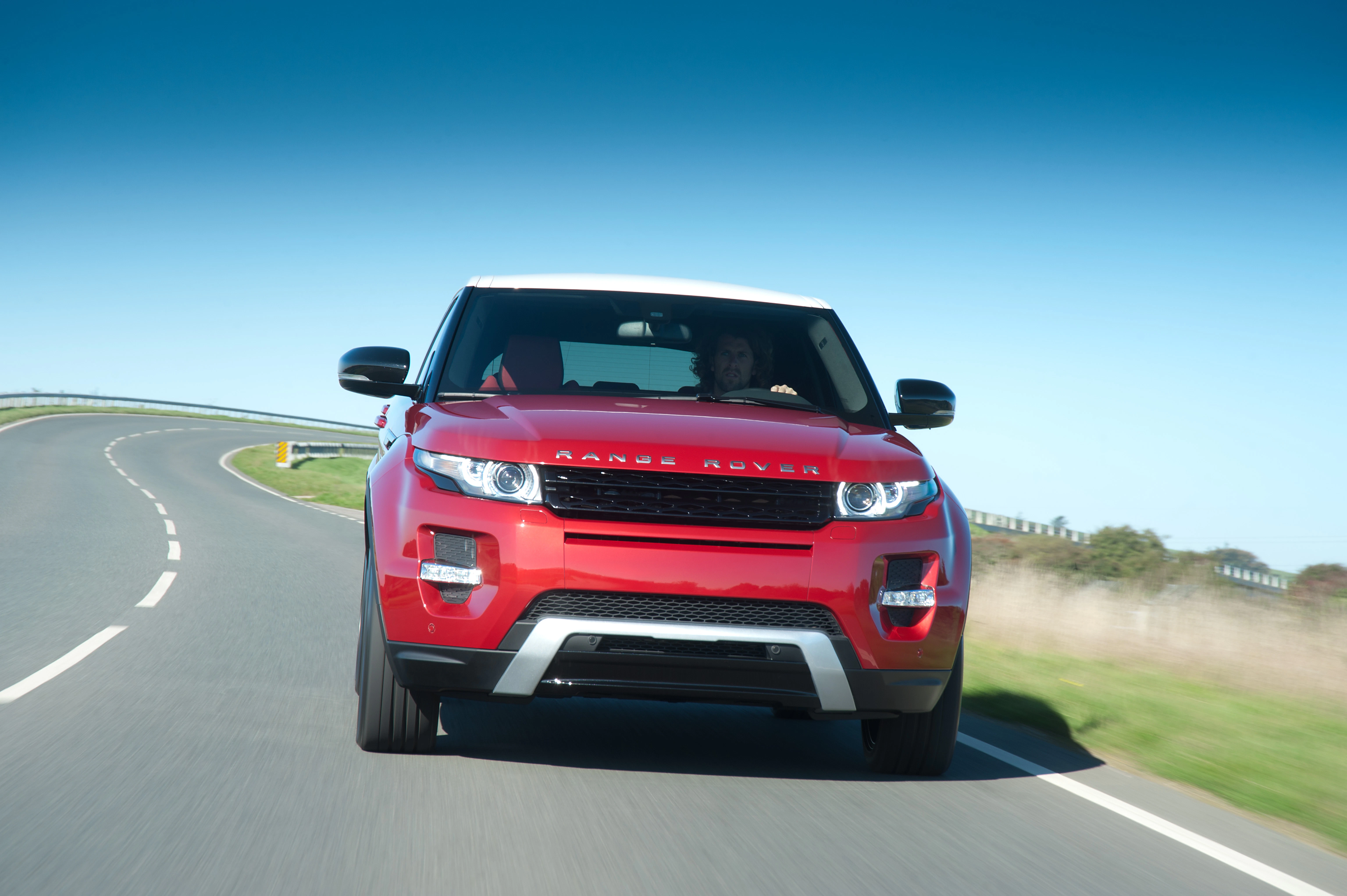 Roter Range Rover fährt auf kurviger Landstraße bei klarem Himmel. Zwei Personen im Auto.