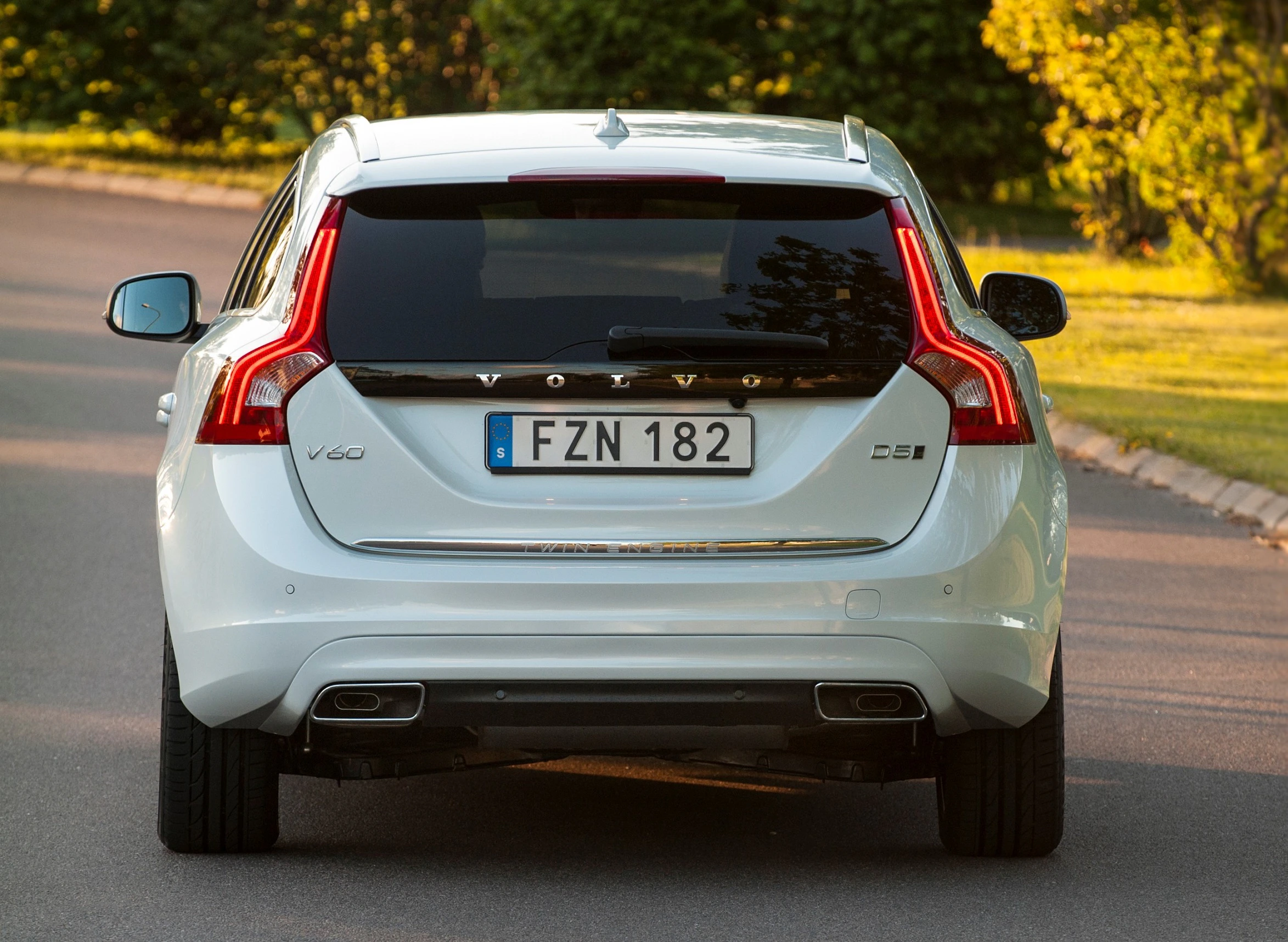 Hinteransicht eines weißen Volvo V60 auf einer Straße, umgeben von grüner Vegetation. nummernschild Lesbar, modernes Rücklichtdesign.