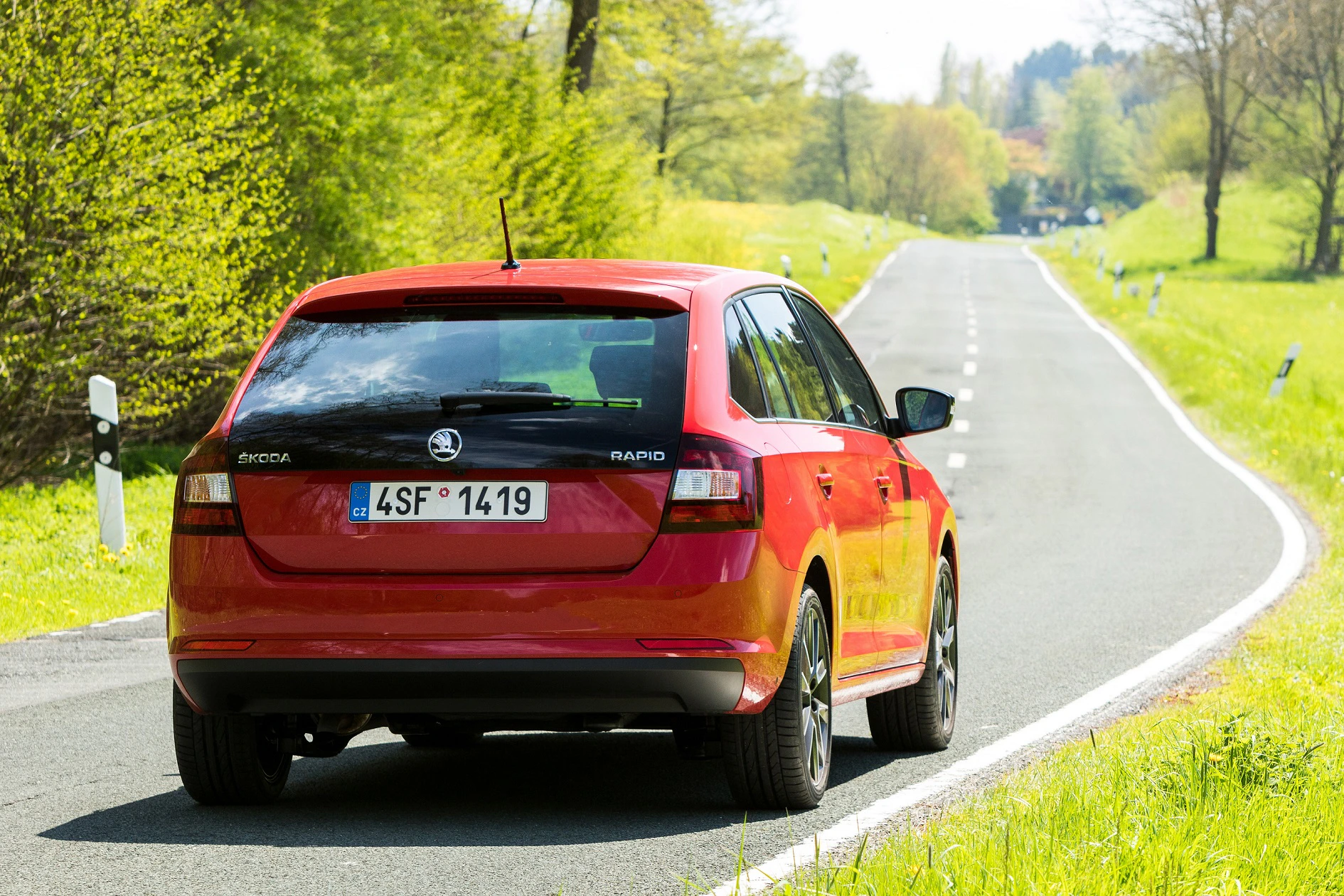 Rotes Auto auf einer ländlichen, kurvigen Straße im Frühling, umgeben von grünen Bäumen und Wiesen.
