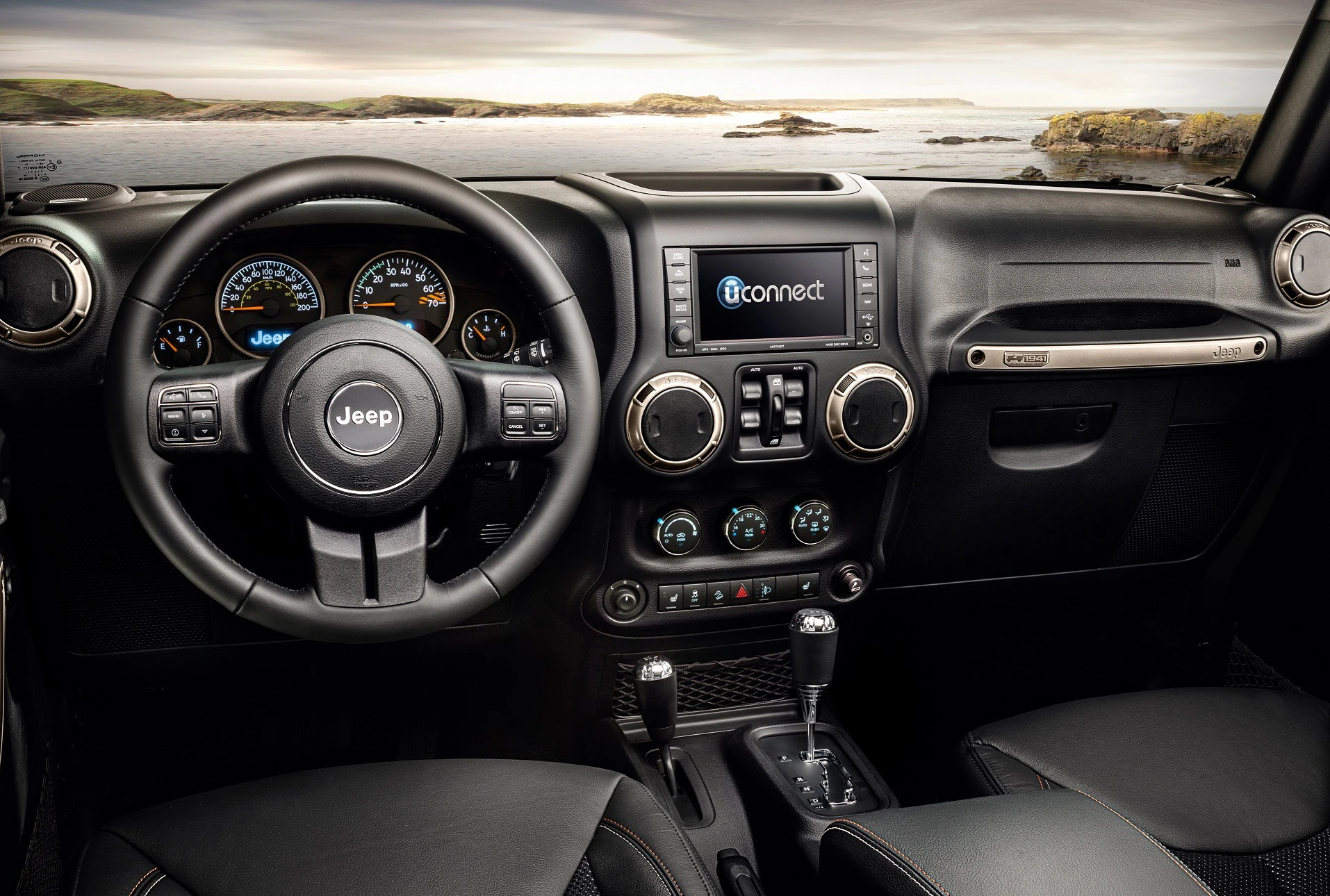 Innenraum eines Jeep mit schwarzem Leder, Lenkrad, Armaturenbrett mit Uconnect-System und Blick auf eine Küstenlandschaft im Hintergrund.