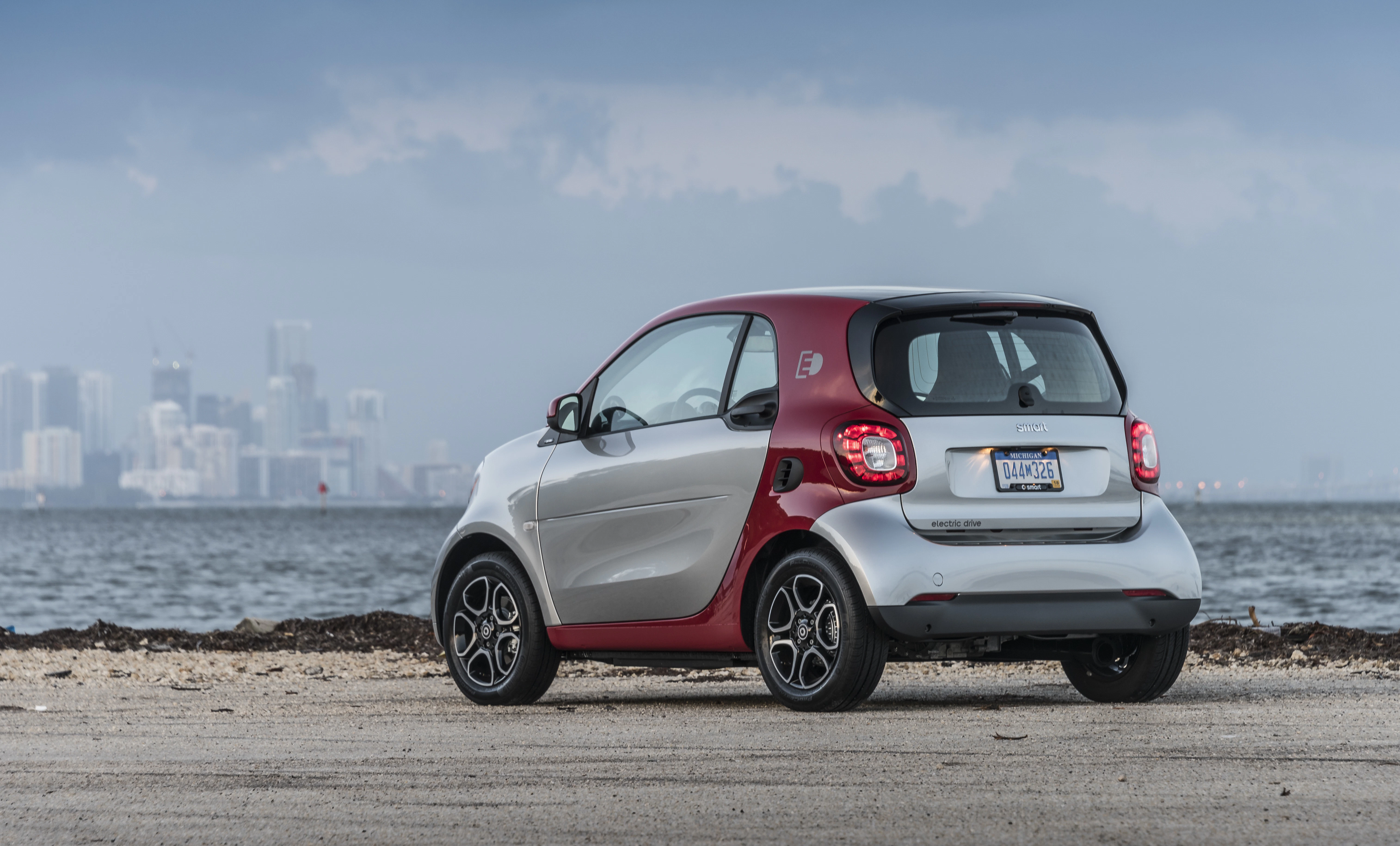 Kleiner zweifarbiger Smart auf einem Strandparkplatz mit Skyline im Hintergrund, bewölkter Himmel.