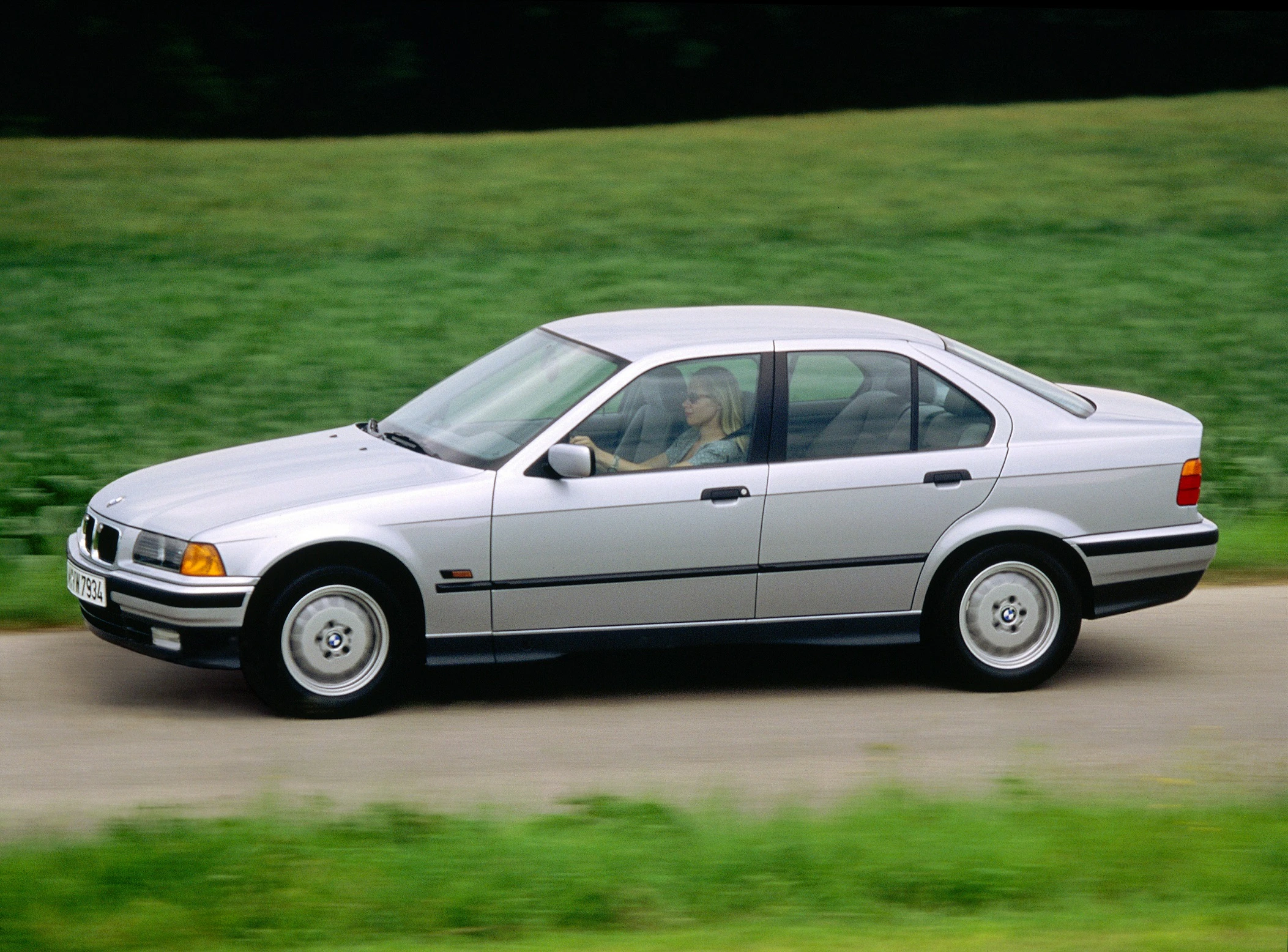 Silberner BMW 3er E36 Limousine fährt auf einer Landstraße, grüne Wiesen im Hintergrund, seitliche Ansicht.