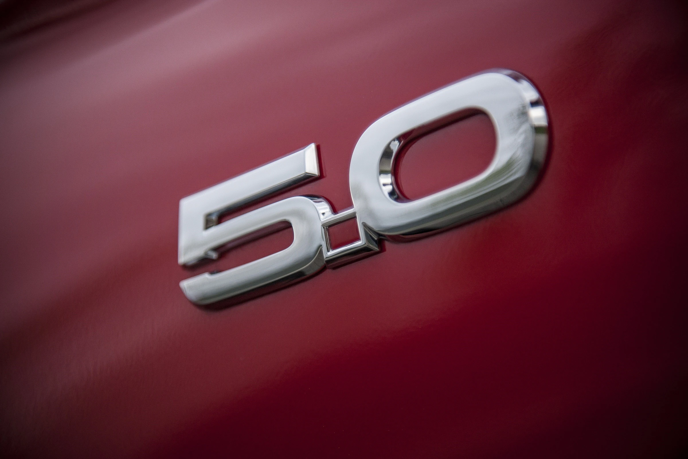 Nahaufnahme eines glänzenden, silbernen "5.0" Emblems auf einem glänzenden roten Auto.