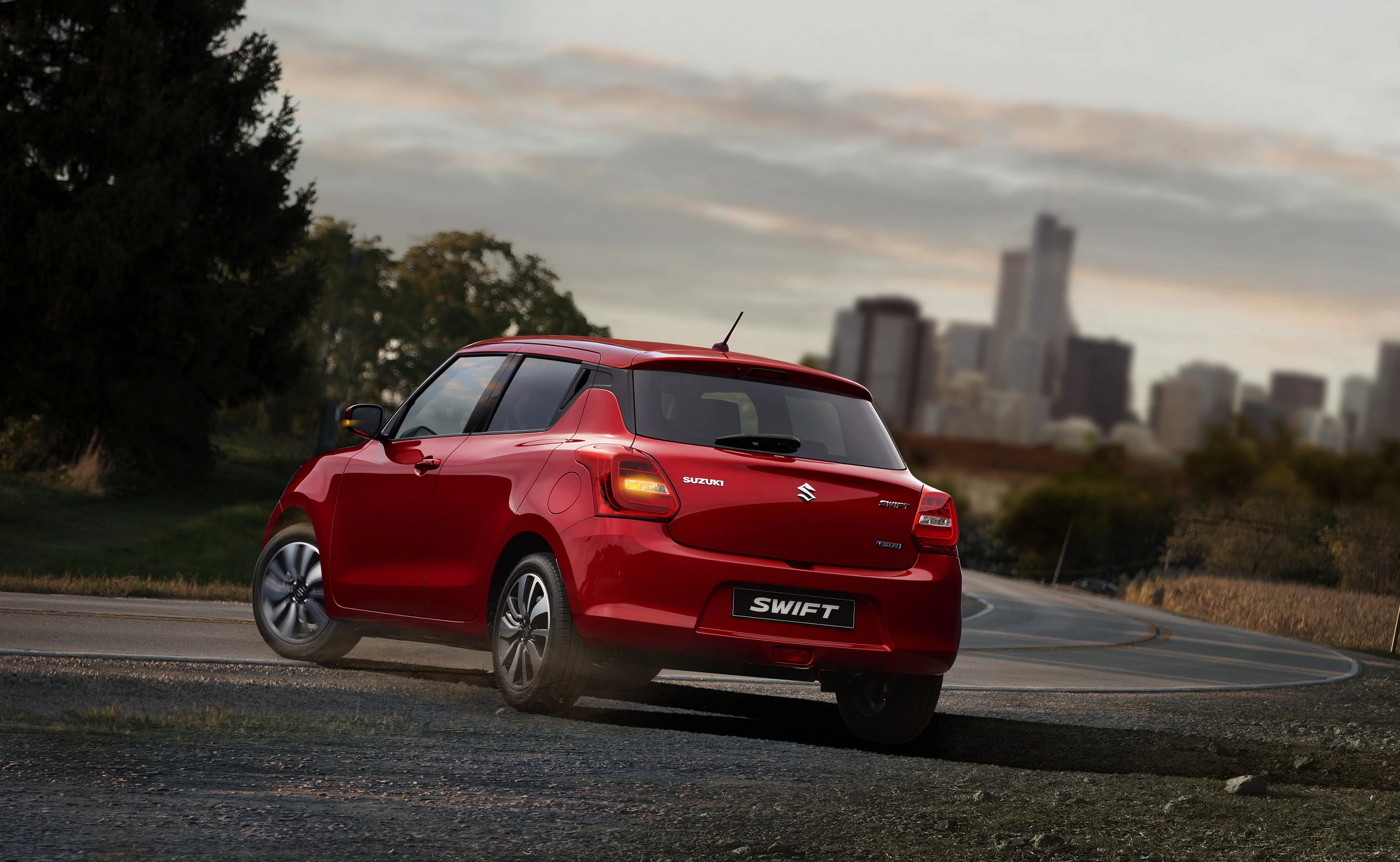 Roter Suzuki Swift fährt auf einer Straße, im Hintergrund eine Stadtlandschaft mit Wolkenkratzern bei Sonnenuntergang.