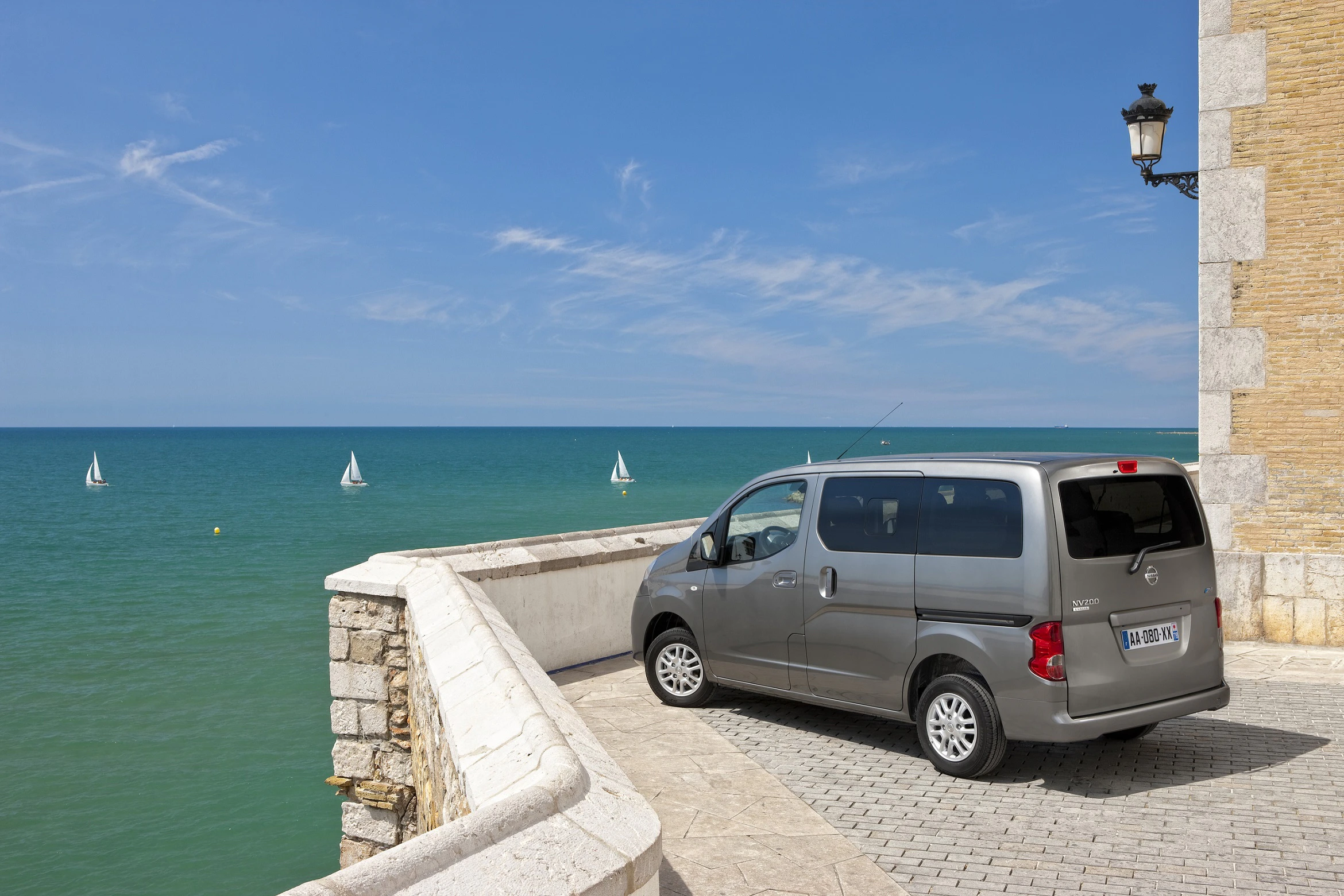 Silberner Van geparkt auf einer Steinpromenade mit Blick auf das Meer und mehrere Segelboote; klare blaue Himmel und eine antike Laterne.