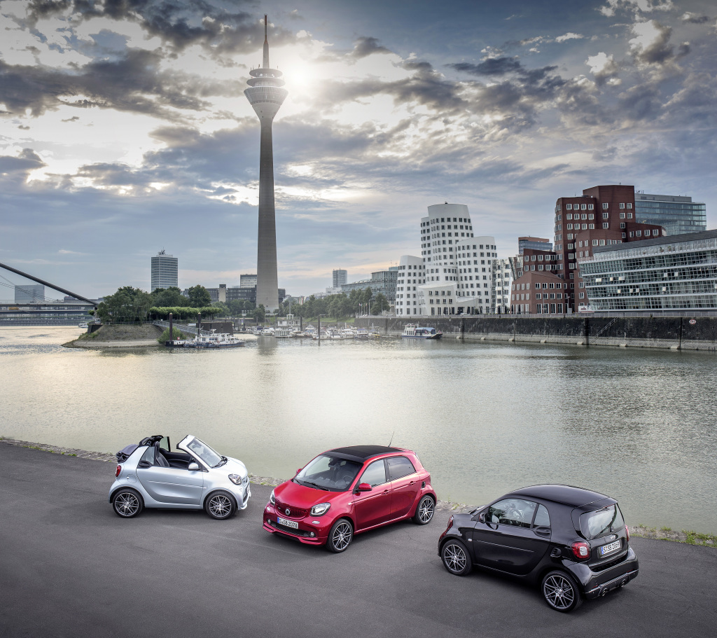 Drei bunte Smarts stehen vor dem Rheinturm und modernen Gebäuden am Rheinufer in Düsseldorf bei bewölktem Himmel.