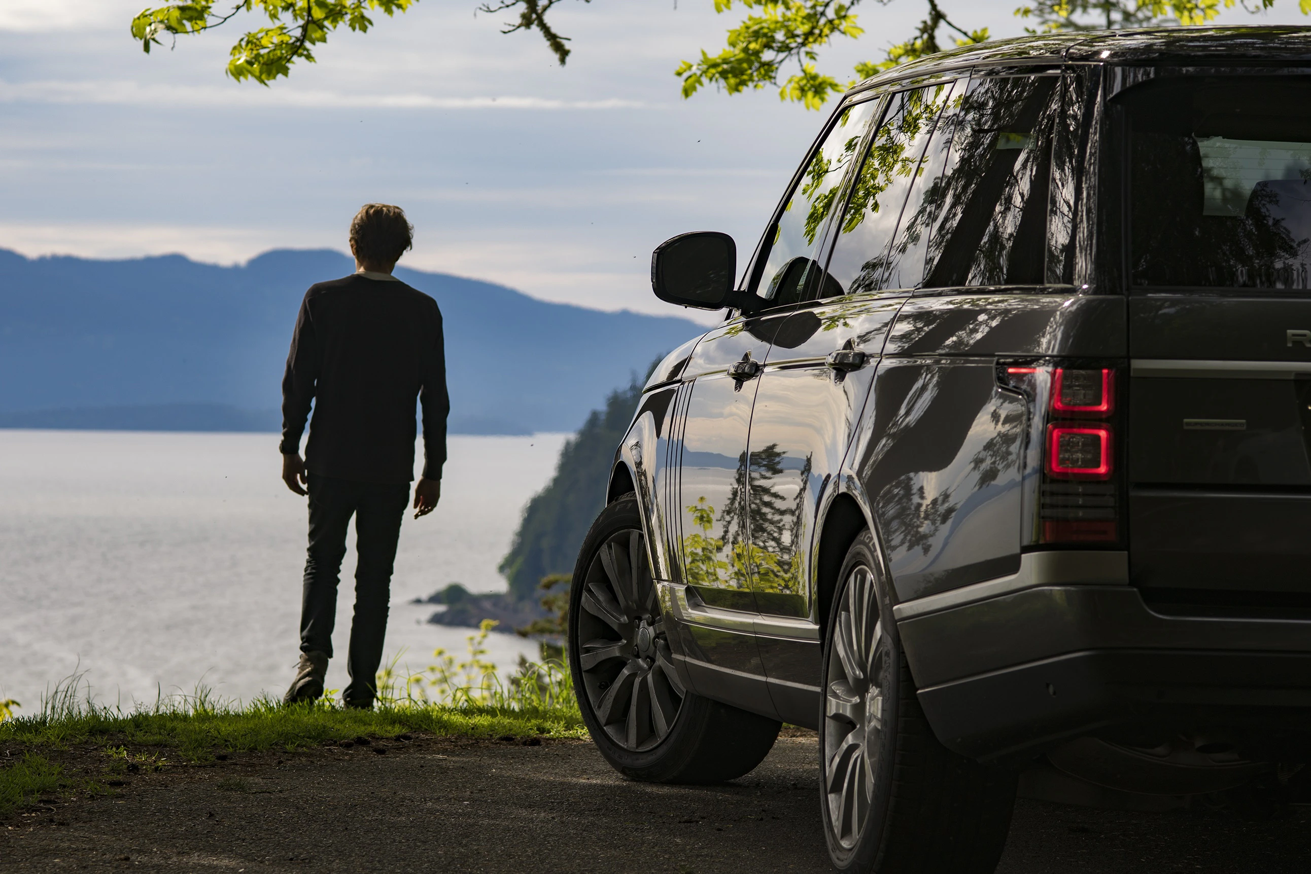 Mann steht am Wasser und blickt auf Landschaft; im Vordergrund ein geparkter SUV bei bewölktem Himmel, Bäume im Hintergrund.