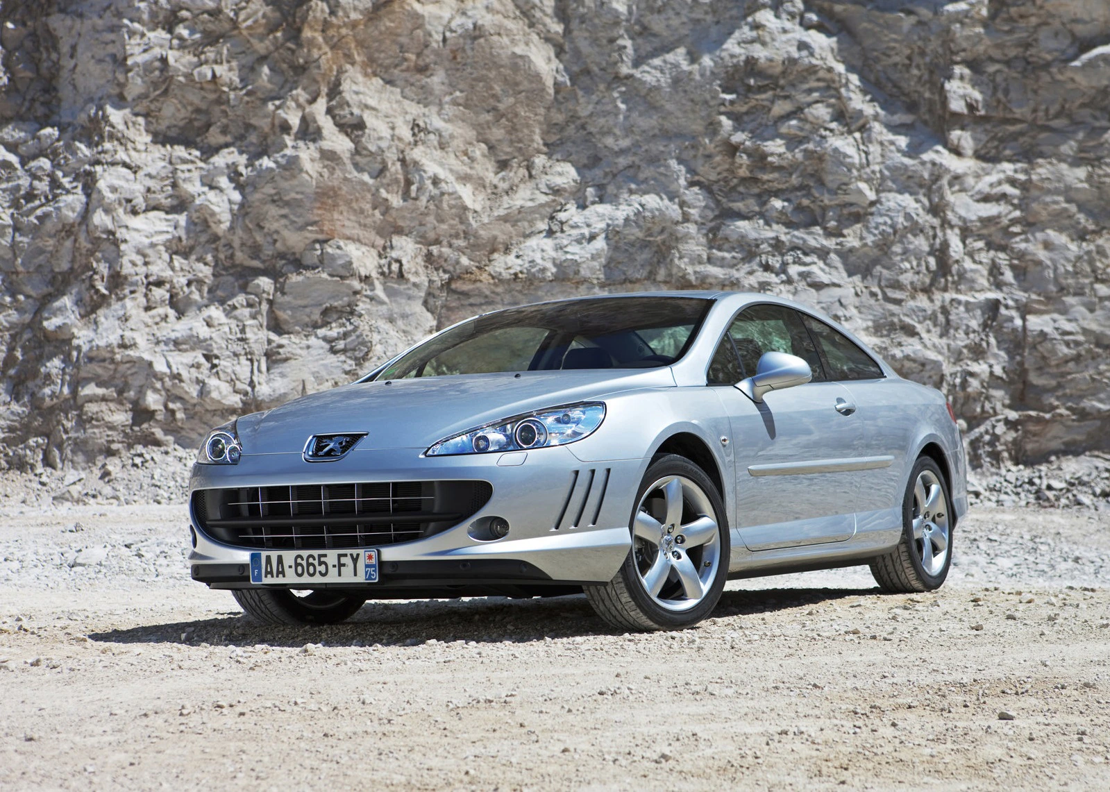 Silberner Peugeot 407 Coupé vor einer felsigen Hintergrundkulisse, geparkt auf sandigem Untergrund.