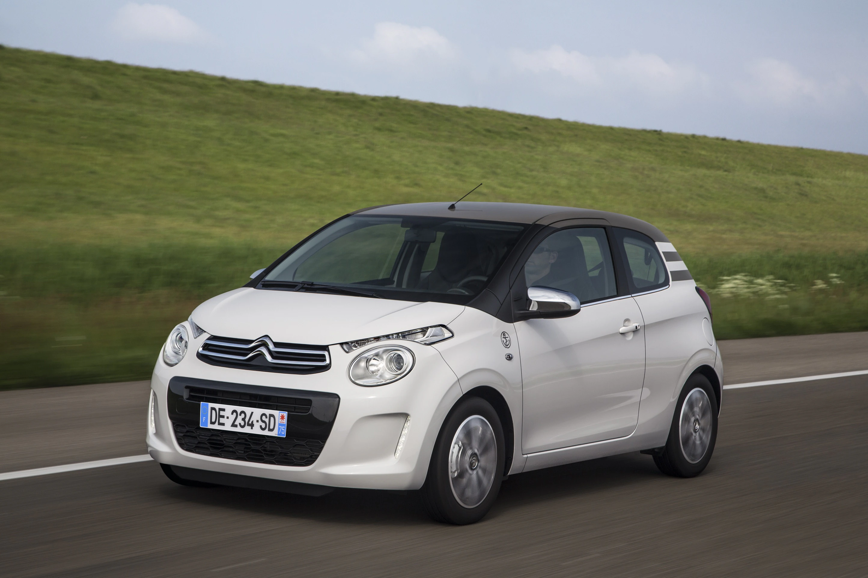 Weißer Citroën C1 fährt auf ländlicher Straße entlang grüner Wiesen, bewölkter Himmel im Hintergrund.