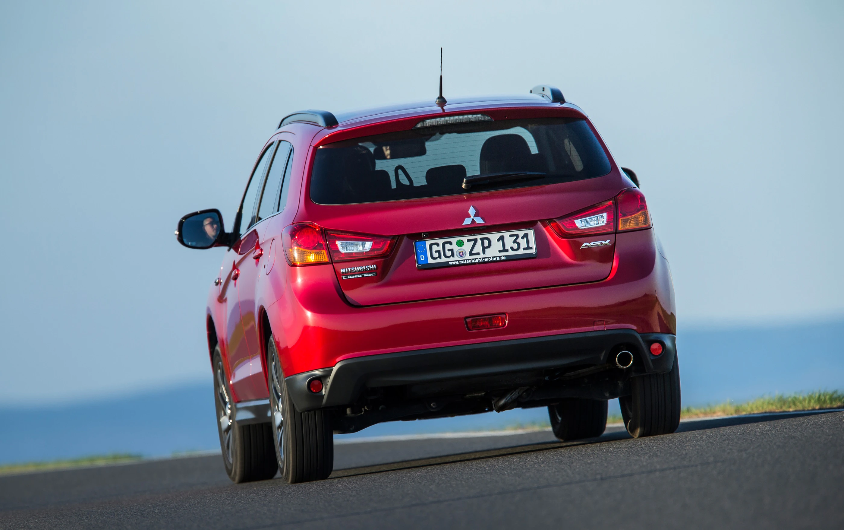 Roter Mitsubishi ASX fährt auf einer Straße bei blauem Himmel, Heckansicht mit deutschem Kennzeichen sichtbar.