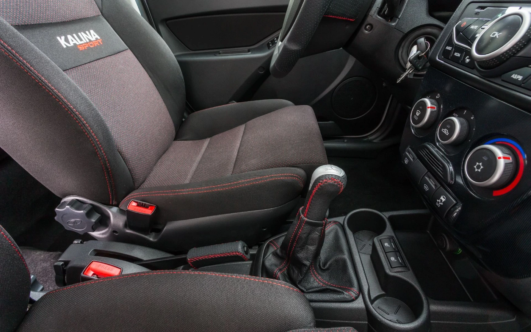 Innenraum eines Autos mit schwarzen Sportsitzen, roten Nähten und einem manuellen Getriebe. Lenkrad und Armaturenbrett mit Bedienelementen.