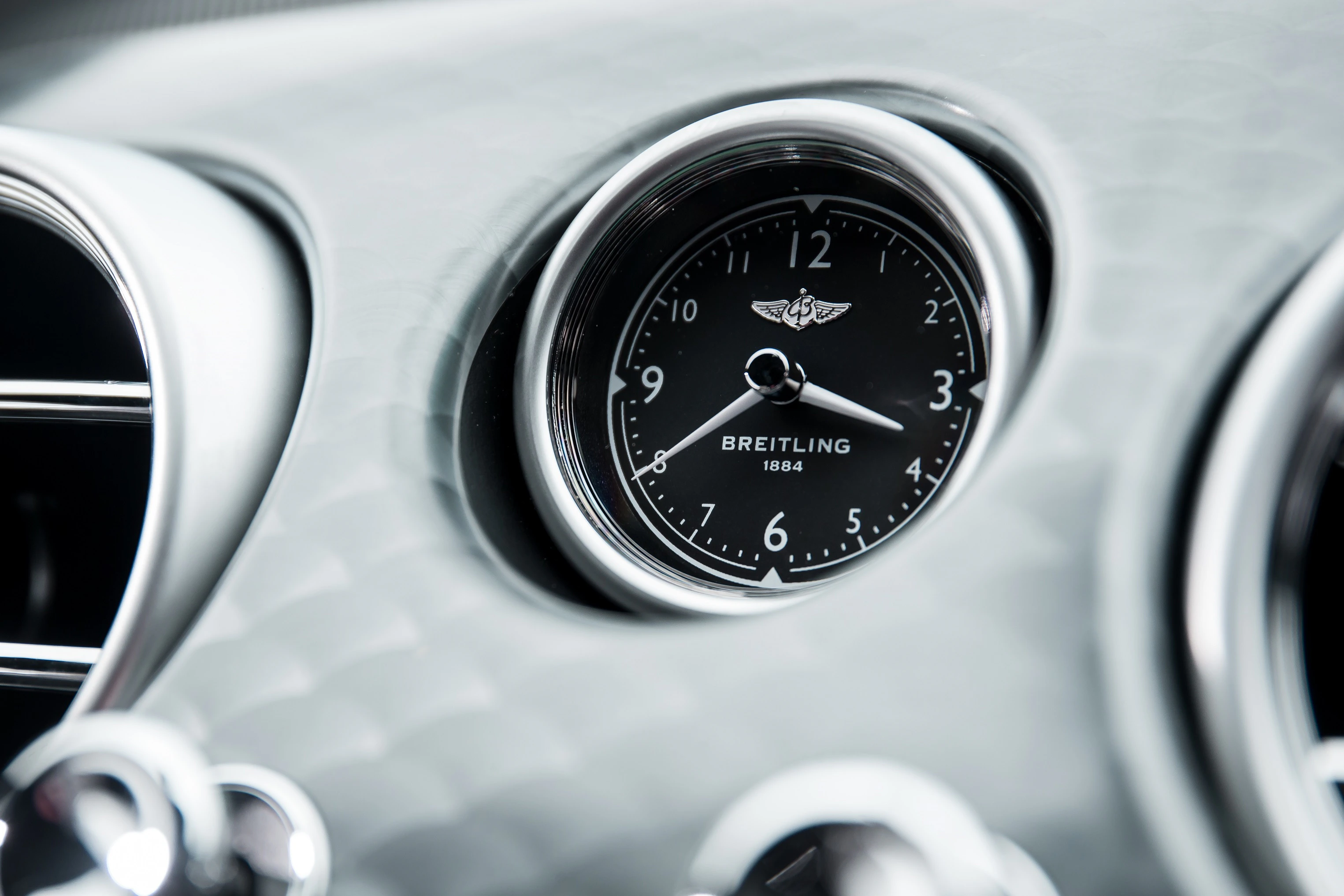 Nahaufnahme einer analogen Uhr von Breitling, eingebaut im Armaturenbrett eines Autos mit einem eleganten, silbernen Zifferblatt.