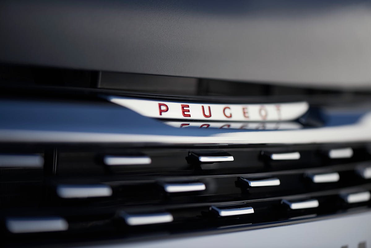 Nahaufnahme eines Peugeot-Logos auf einem Fahrzeuggrill mit modernen, dunklen Linien und spiegelnder Oberfläche.