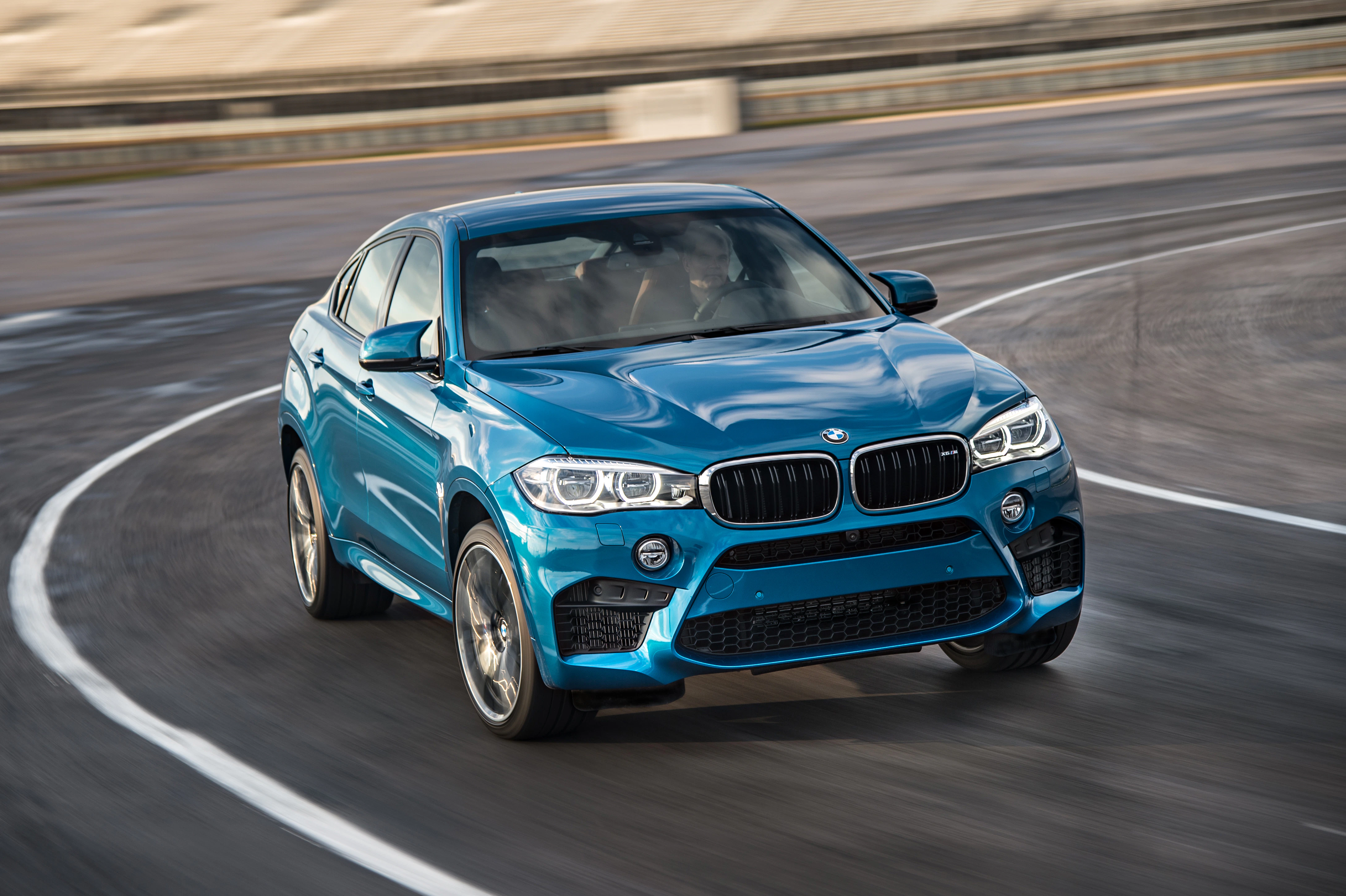 Blauer BMW X6 fährt schnell auf einer leeren, kurvigen Rennstrecke. Dynamische Bewegung, sportliches Design und klare Himmel im Hintergrund.