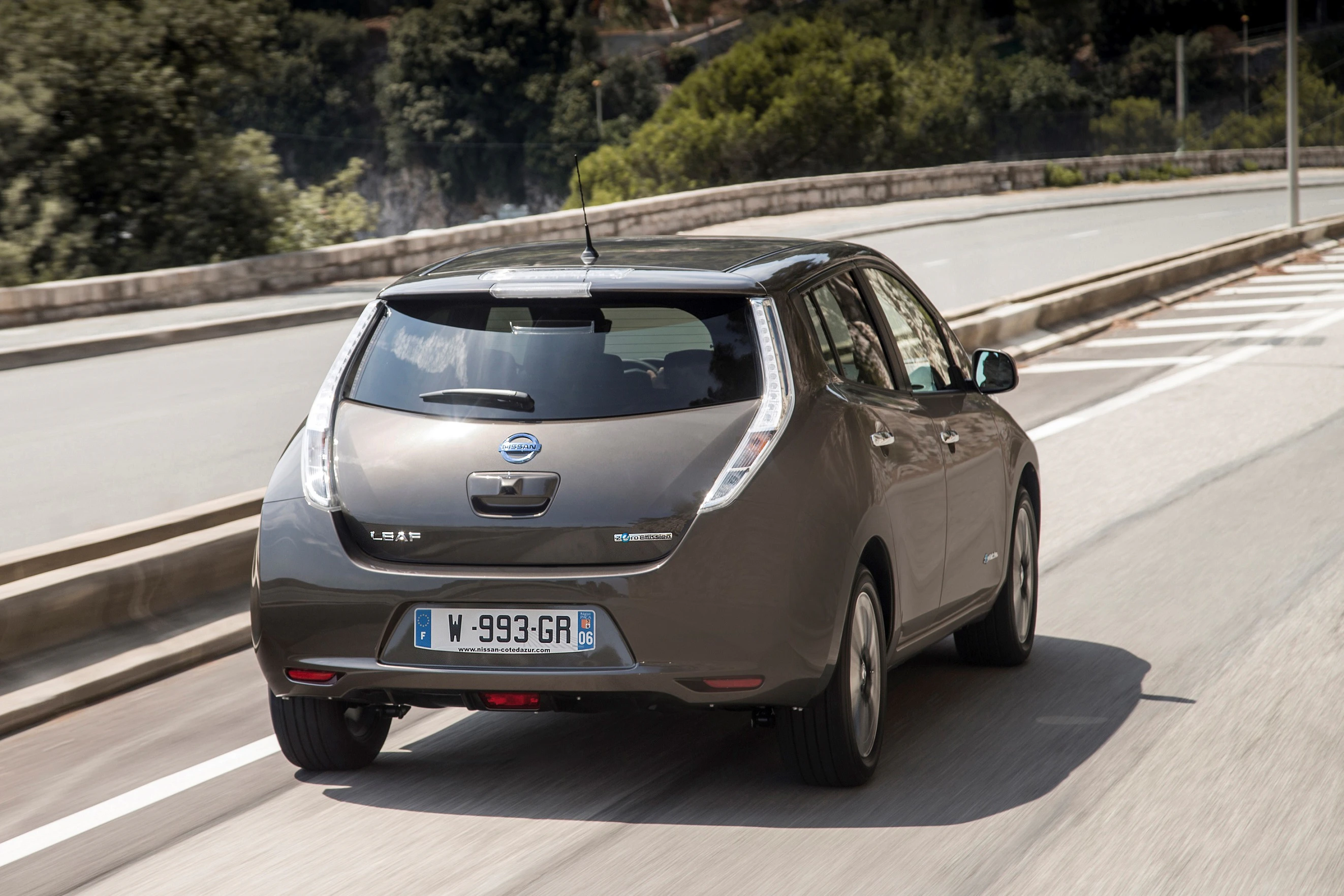 Ein grauer Nissan Leaf fährt auf einer Straße mit Bäumen im Hintergrund.