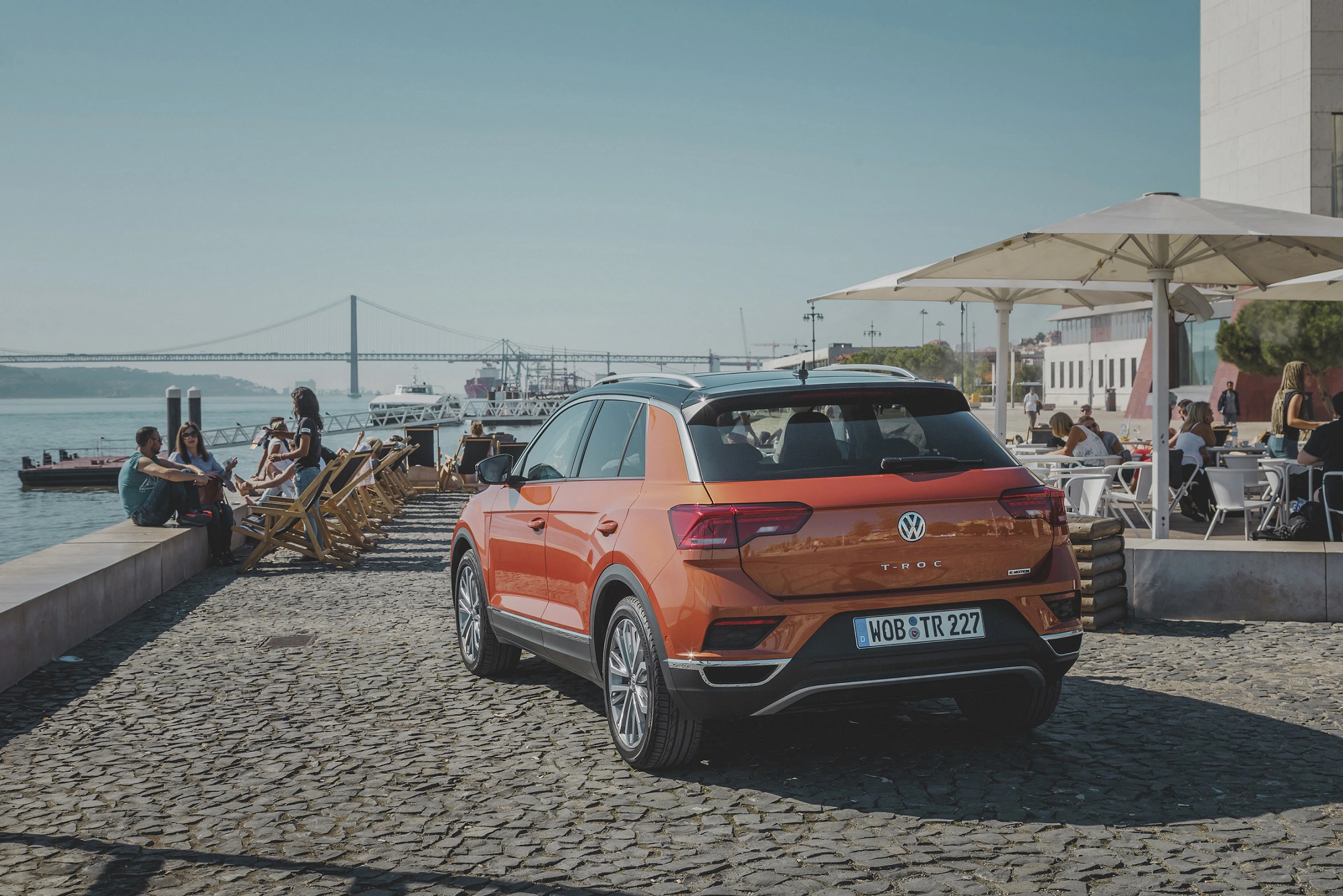 Ein orangefarbener VW T-Roc parkt auf einer gepflasterten Uferpromenade. Im Hintergrund entspannen Menschen in Cafés und genießen die Aussicht auf den Fluss.