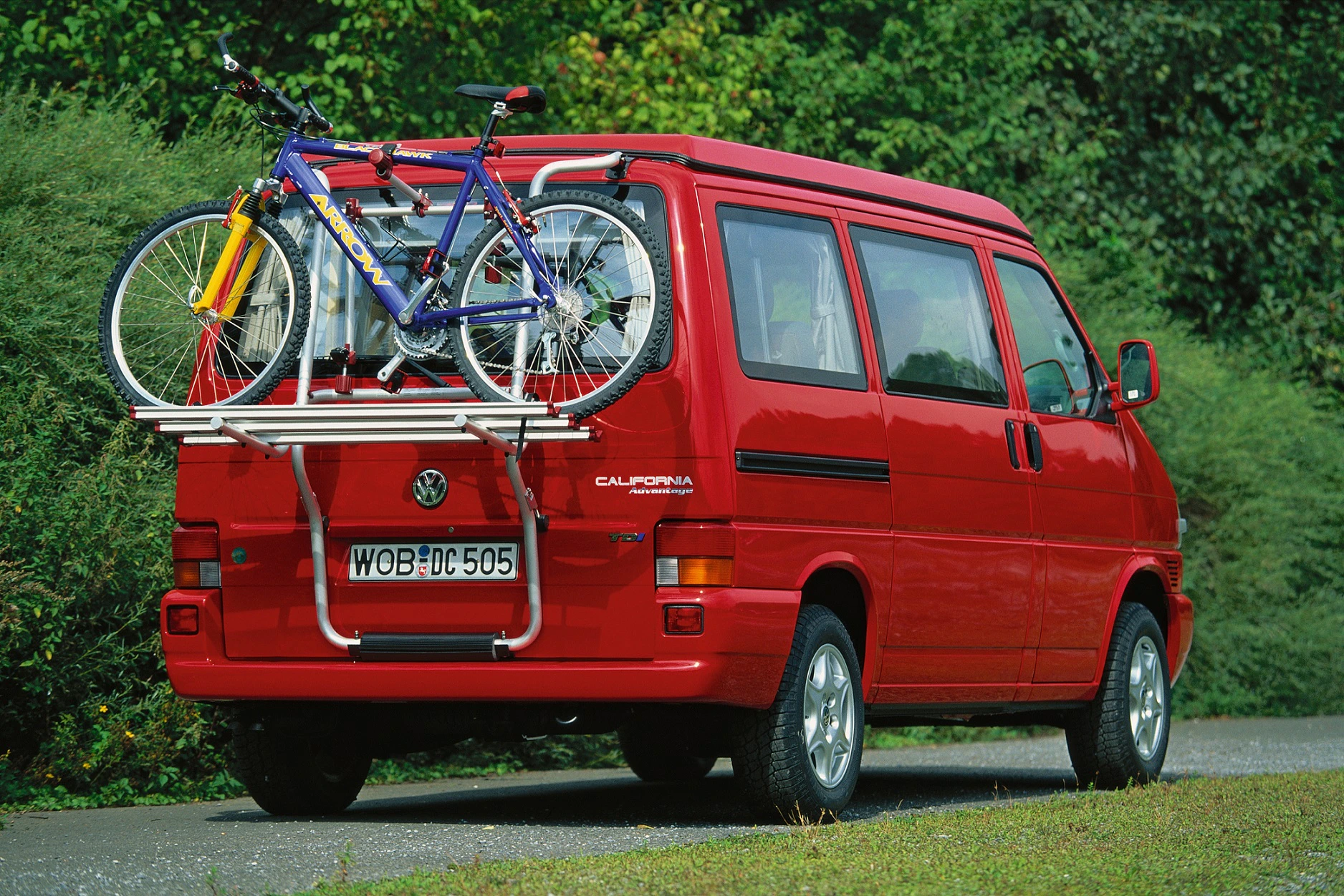 Roter VW-Bus mit auf dem Heckträger montierten Fahrrädern, auf einer Straße von Bäumen und Büschen flankiert.