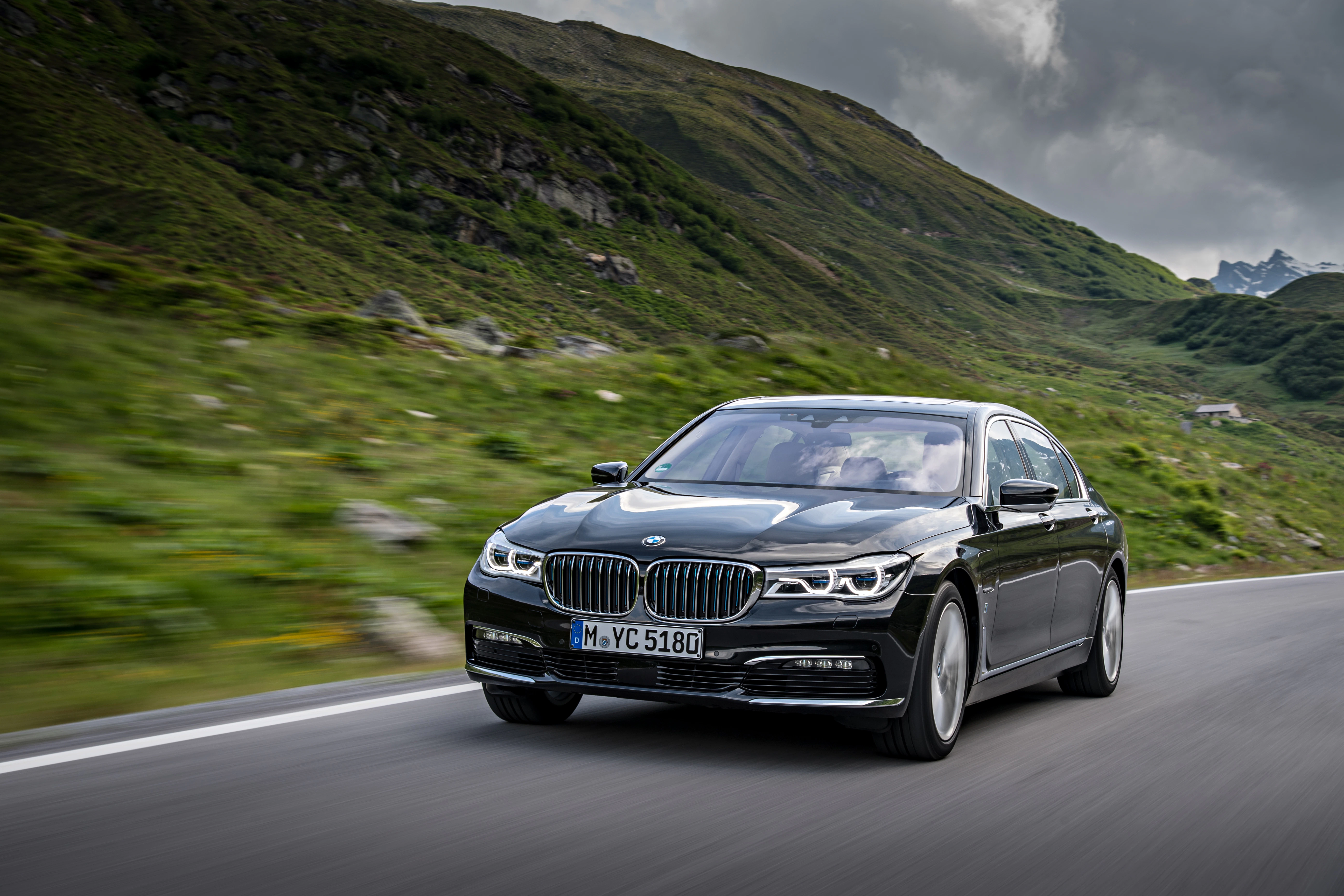 Schwarzer BMW fährt auf einer Landstraße durch eine grüne, bergige Landschaft unter bewölktem Himmel.