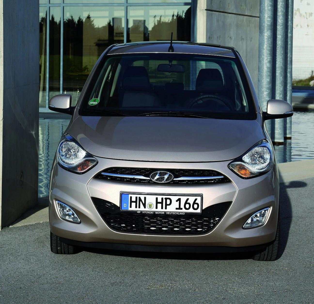 Silberfarbener Hyundai i10, frontal, vor modernem Gebäude mit Glasfassade und Wasserbecken, Tageslicht.