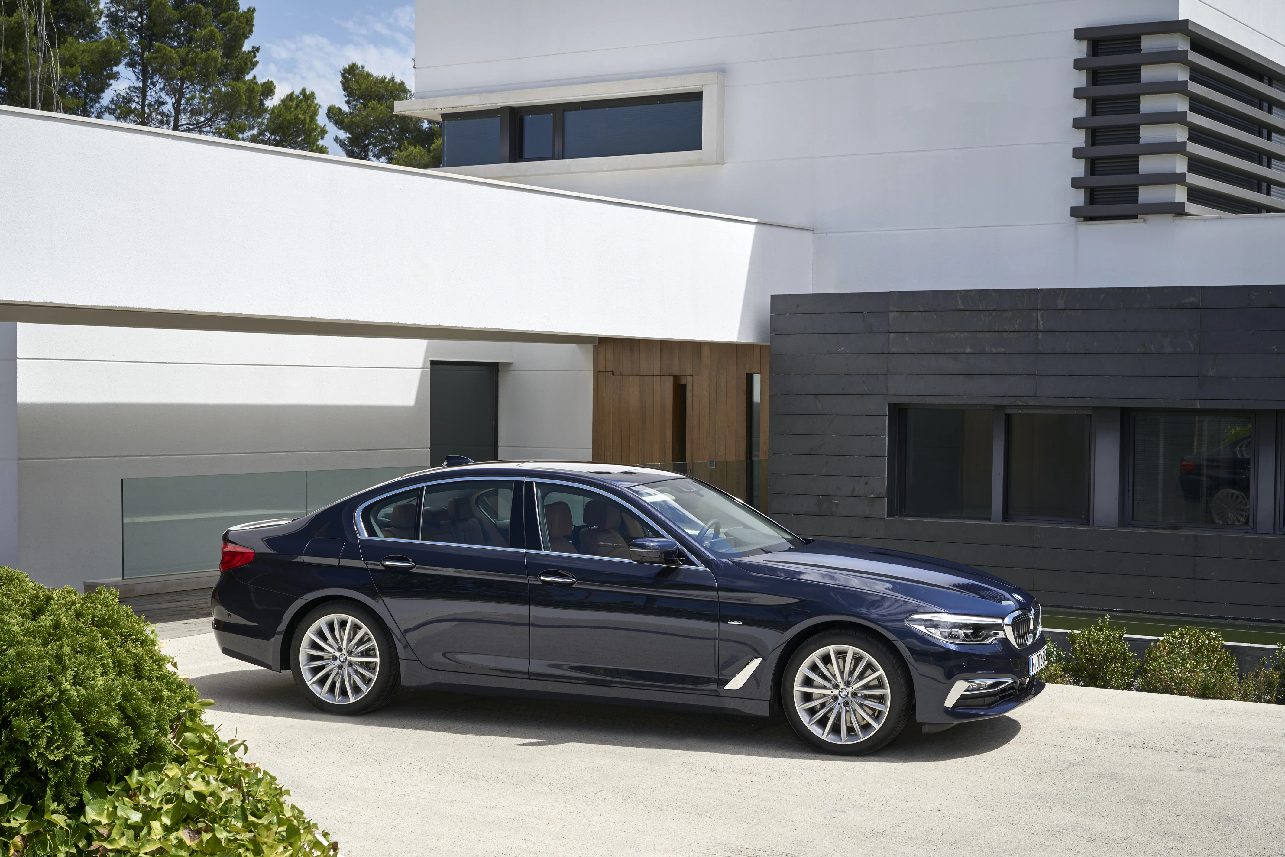Dunkelblauer Luxuslimousine vor einem modernen, weißen Gebäude mit großen Fenstern und grünen Büschen im Vordergrund.