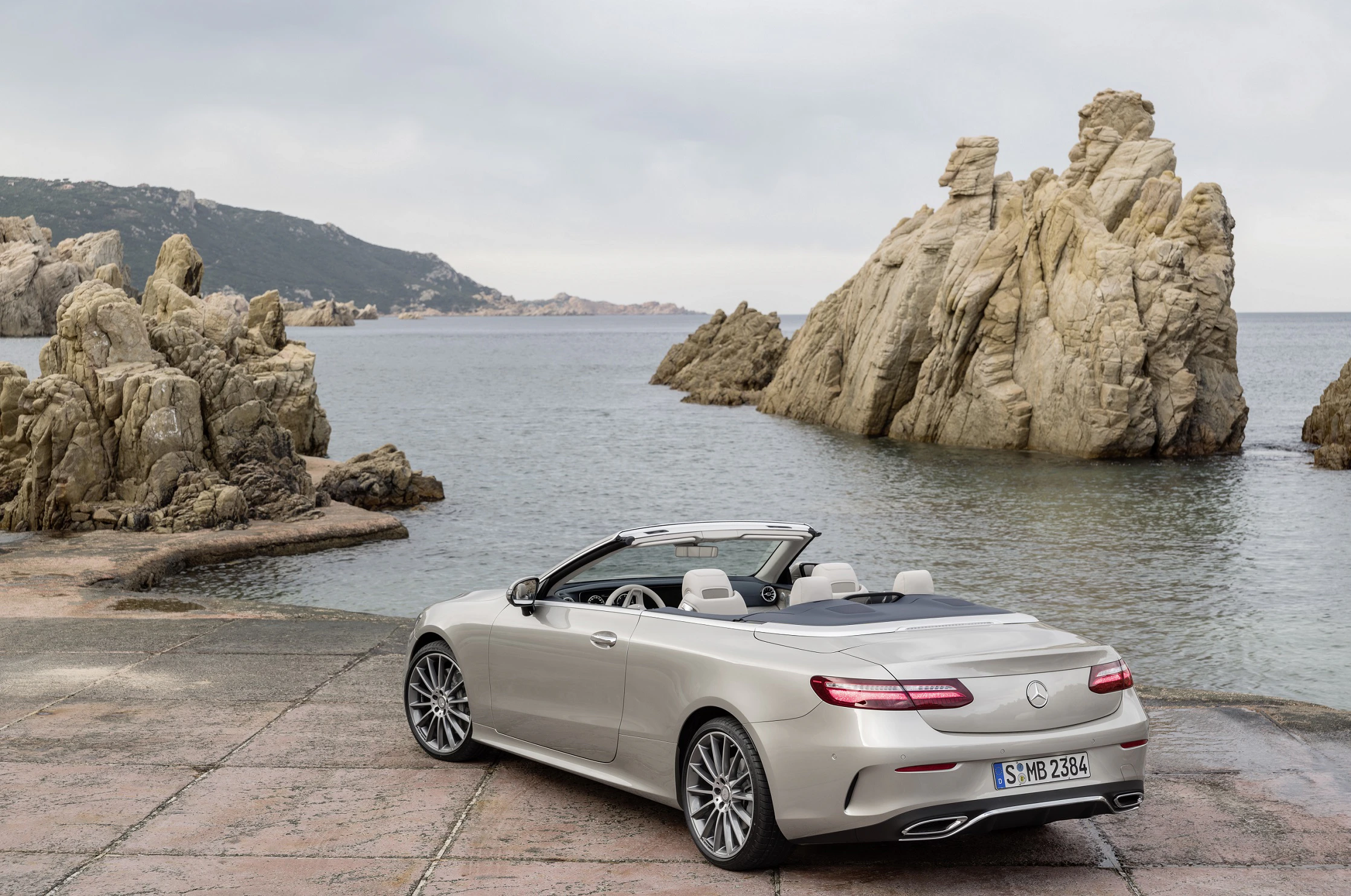 Silbernes Cabrio parkt vor steiniger Küstenlandschaft am Meer unter bewölktem Himmel.