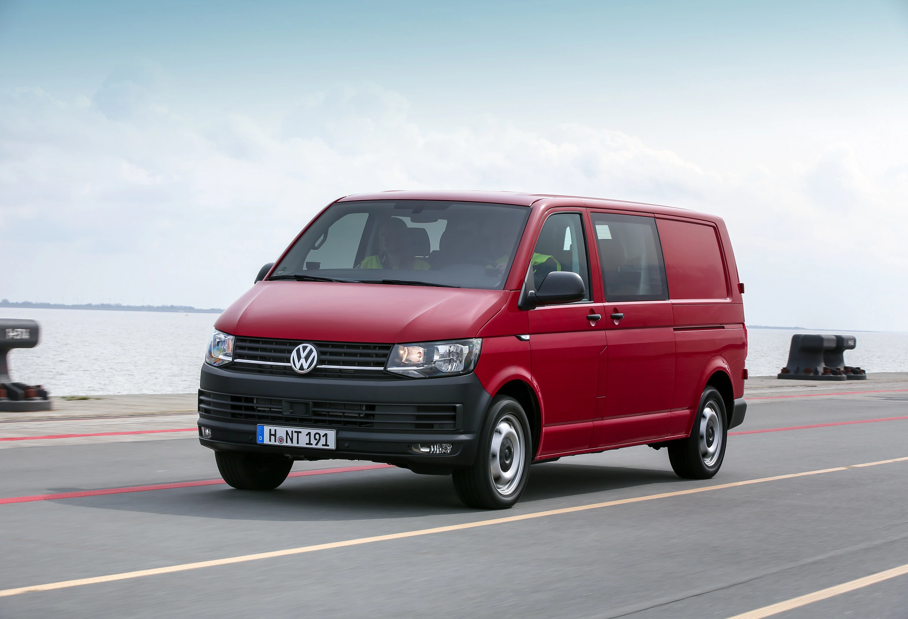 Roter VW Transporter fährt auf einer Küstenstraße entlang, im Hintergrund ist das Meer zu sehen.