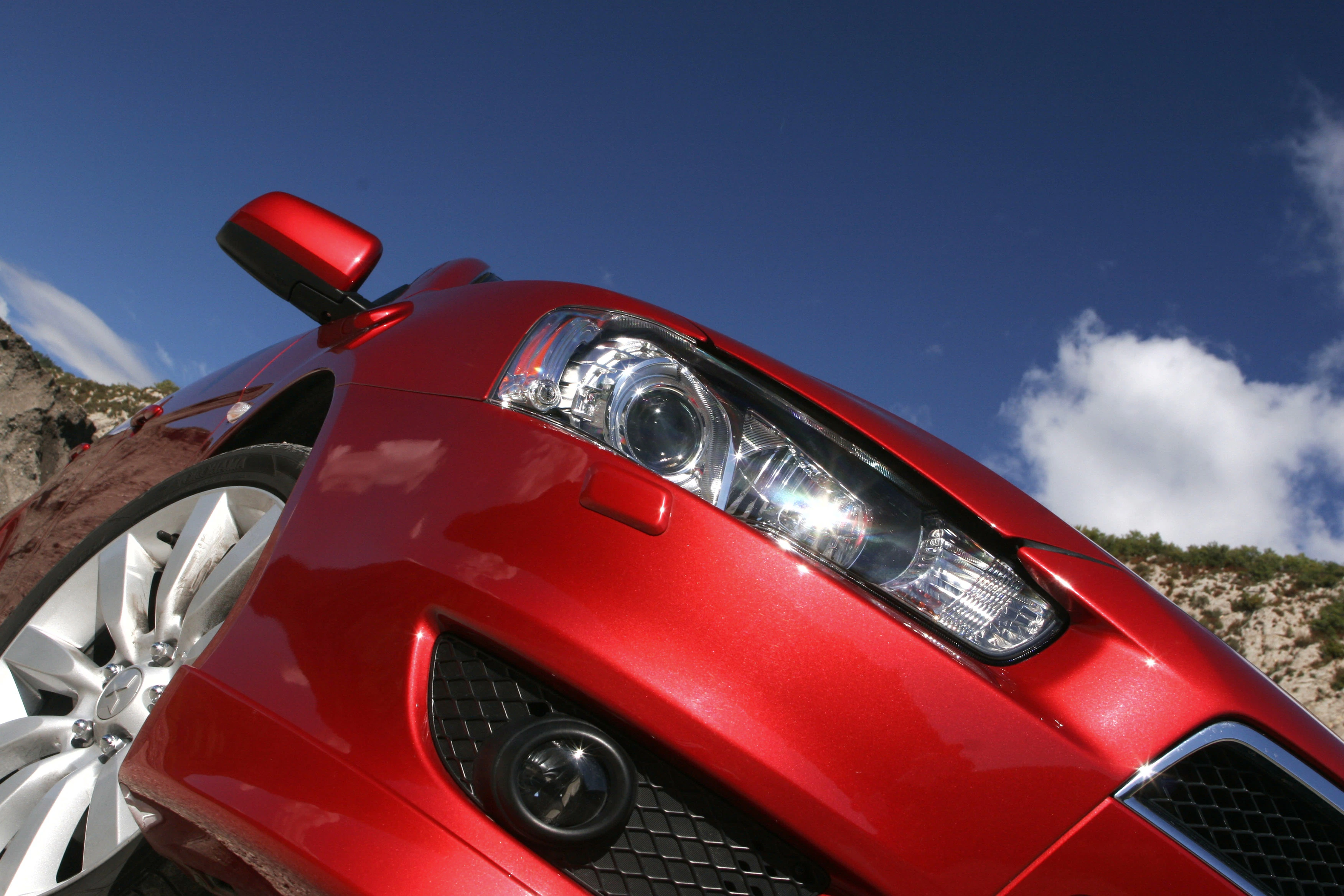 Roter Sportwagen im sonnigen Freien, von unten fotografiert, mit strahlenden Scheinwerfern und blauem Himmel im Hintergrund.